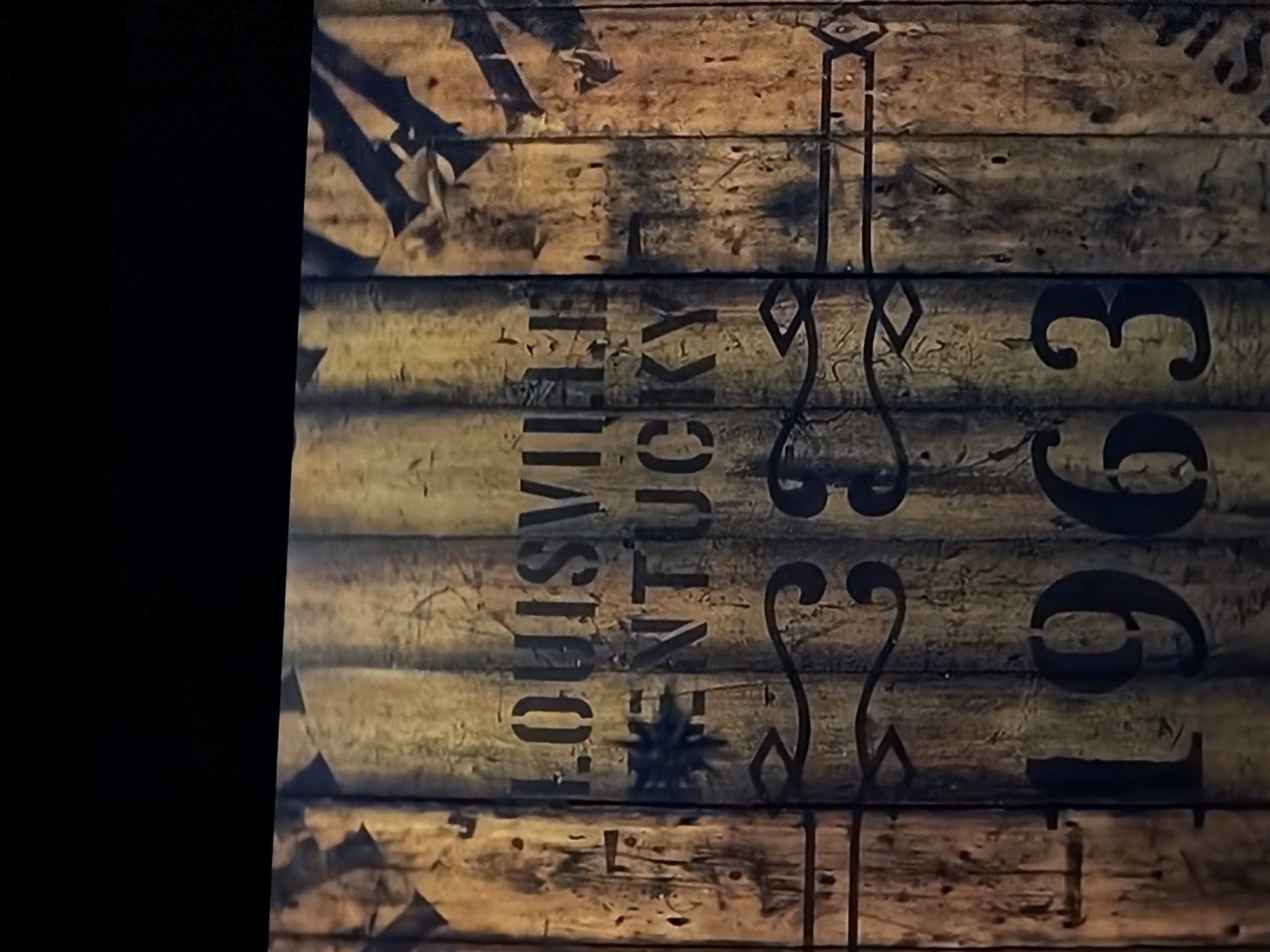 The image presents a close-up, vertically oriented view of a rustic wooden surface, likely planks or staves from a crate, barrel, or sign. The wood exhibits a textured, aged appearance with visible grain and varying shades of light brown, yellow, and darker brown, suggesting weathering or a distressed finish. The left portion of the image is completely dark, indicating poor lighting or an obscured area.
Prominently displayed in black stencil or painted lettering are words and numerals. Vertically oriented along the center, the text "LOUISVILLE, KY" is clearly visible, with "LOUISVILLE" stacked above "KY". Below "LOUISVILLE," a stylized starburst symbol is present, followed by partially visible letters that form "KENTUCKY," though fragmented across the wooden planks. Running vertically through and alongside the text are ornate, decorative scroll-like elements in black, adding to the vintage aesthetic. To the right of these decorations, the year "1963" is visible, with the numeral '1' stylized as a tall, slender line capped with a diamond shape, followed by the numerals '9', '6', and '3' which also have a decorative, somewhat stencil-like quality.
There is no activity depicted. The scene captures a static object with clear text indicating "Louisville, KY," suggesting a connection to Louisville, Kentucky. The time of day is not discernible, but the lighting appears artificial or localized. The object's weathered state and stylized text imply an antique or heritage item. While the object is presented without a clear wider context, the text directly refers to Louisville, Kentucky.
