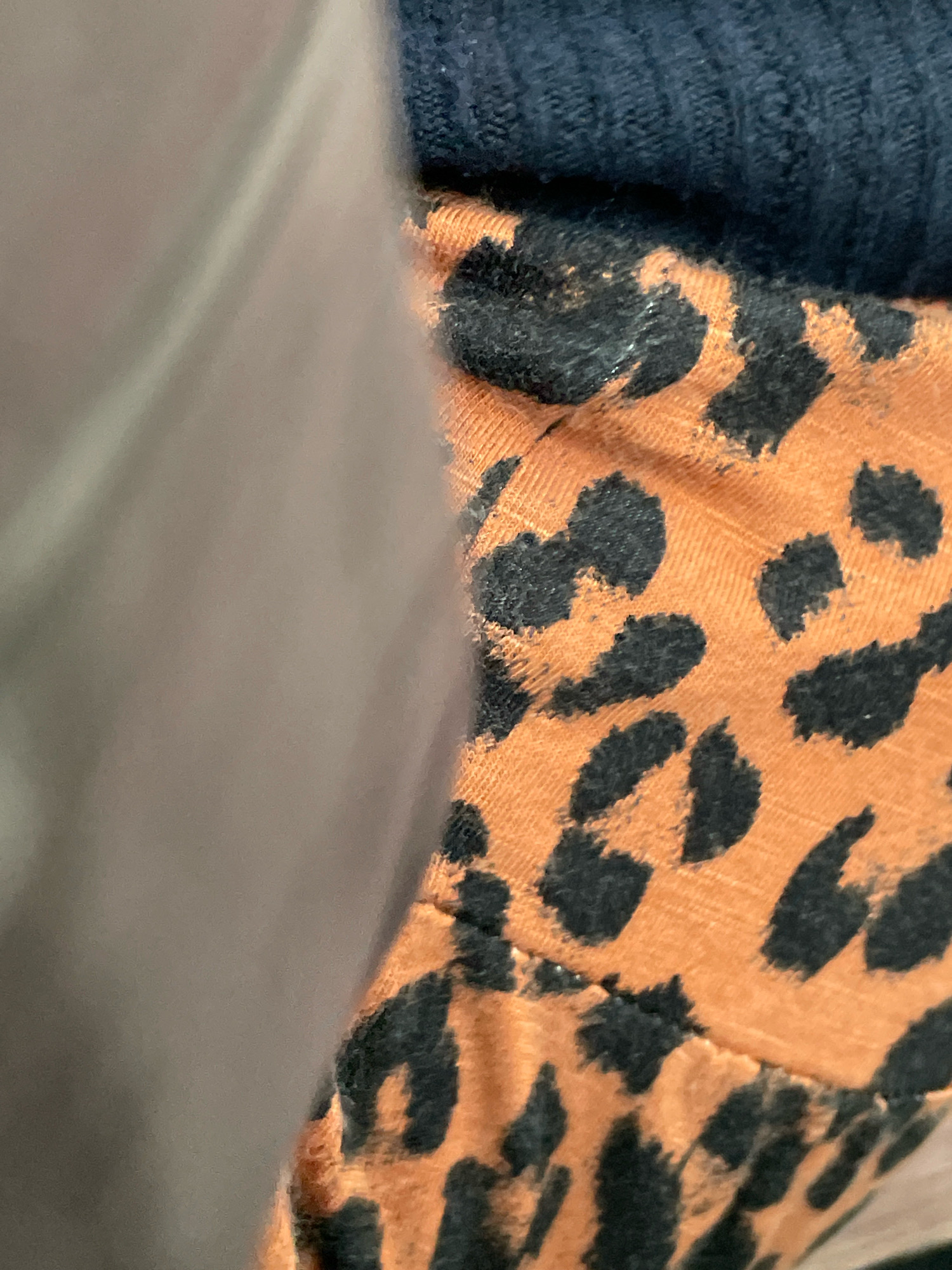 The image is a close-up shot of fabric, displaying a leopard print pattern on an orange background, with a section of dark blue ribbed fabric visible at the top. The leopard spots are irregular in shape and black in color. A blurred, light brown or gray fabric is positioned on the left side, partially obscuring the leopard print. The scene appears to be indoors, and no specific location in Ajah, Nigeria, can be identified from the visual information provided. There are no people or actions depicted in the image.