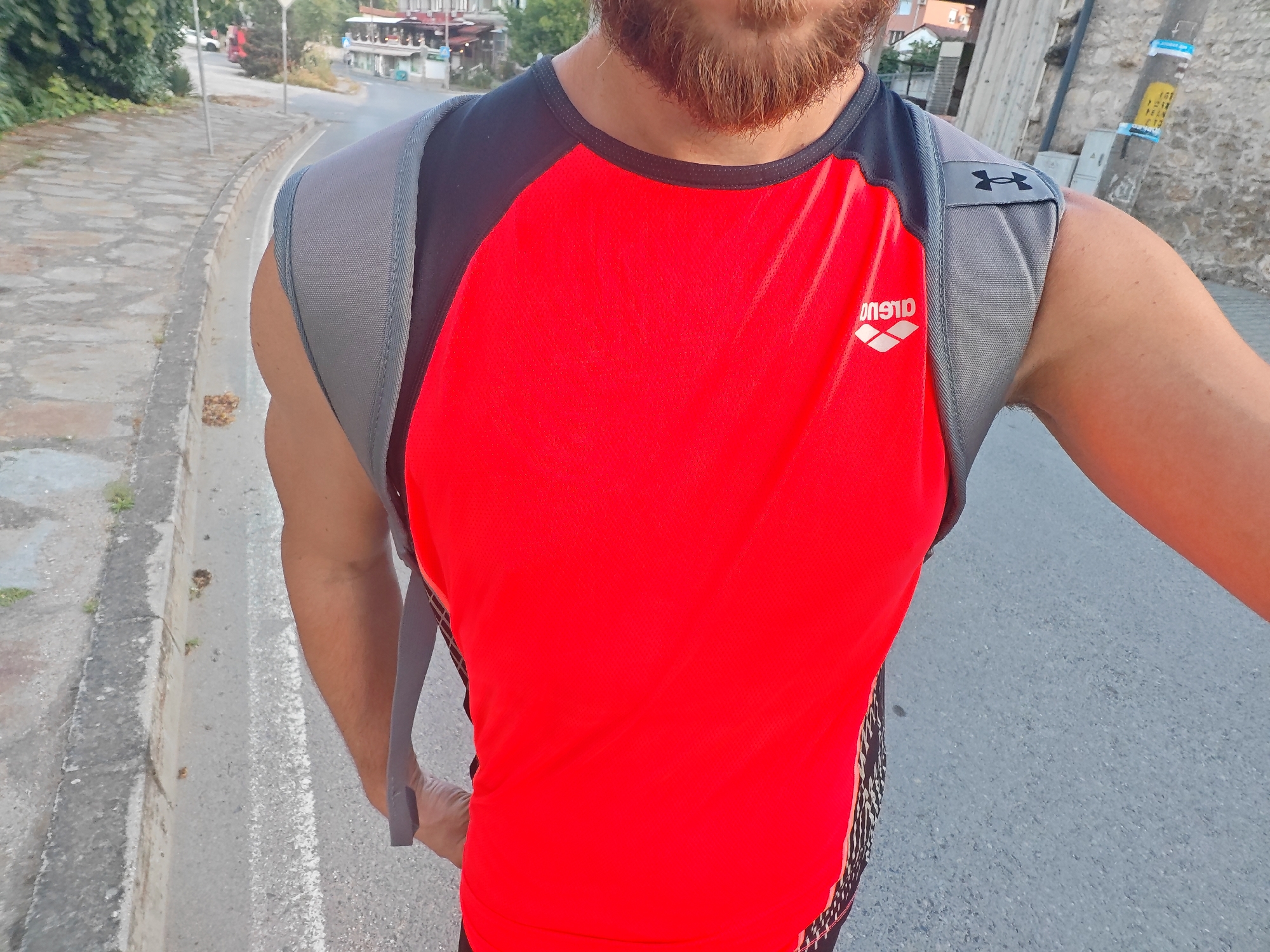 A person is depicted from a first-person, waist-up perspective, standing outdoors on an asphalt road next to a stone-paved sidewalk in the city of Ivaylovgrad, Bulgaria. The individual wears a red sleeveless athletic shirt with black trim around the collar and shoulders, featuring a reversed white "ARENA" logo on the chest. Black and white patterned fabric is visible on the lower sides of the shirt. A grey backpack or vest is worn over the shirt, with a black "Under Armour" logo prominently displayed on the right shoulder strap. The person has a visible beard and light to medium brown skin tone.
To the left of the person, a concrete curb separates the asphalt from the stone-paved sidewalk. Patches of green vegetation are present along the curb. In the mid-ground, a road curves to the left, bordered by dense green trees and bushes. In the background on the left, several buildings with light-colored facades and terracotta roofs are visible, characteristic of a town setting. A white vehicle and a red truck are partially visible further down the road. To the right of the person, a tall, grey concrete or stone wall runs parallel to the road. A utility pole with multiple attached yellow and blue advertisements or signs stands near the wall, with a small grey utility box at its base. The scene is illuminated by natural daylight, suggesting late afternoon or early morning.