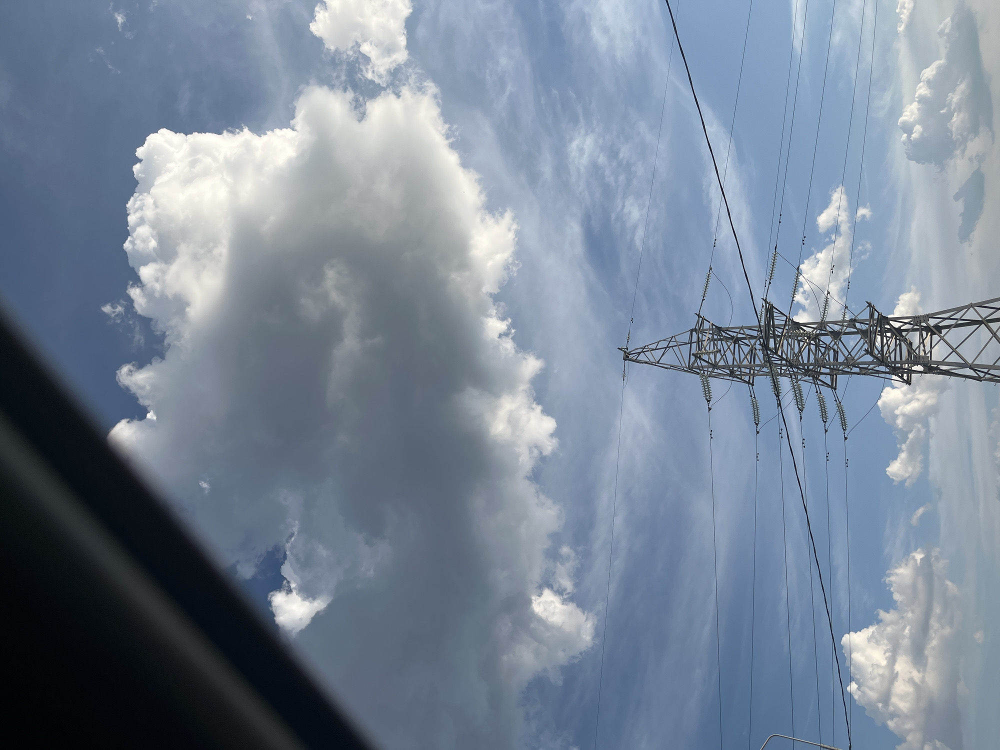 A tall, metal electricity pylon stands prominently in the center of the frame, its lattice structure silhouetted against a bright blue sky. Multiple power lines extend horizontally from the pylon, disappearing out of frame. Scattered white clouds drift across the sky, some large and fluffy, others thin and wispy. A dark, diagonal shadow cuts across the top left of the image, obscuring a portion of the sky and clouds. The scene appears to be located in an outdoor, possibly rural or semi-urban, area.
