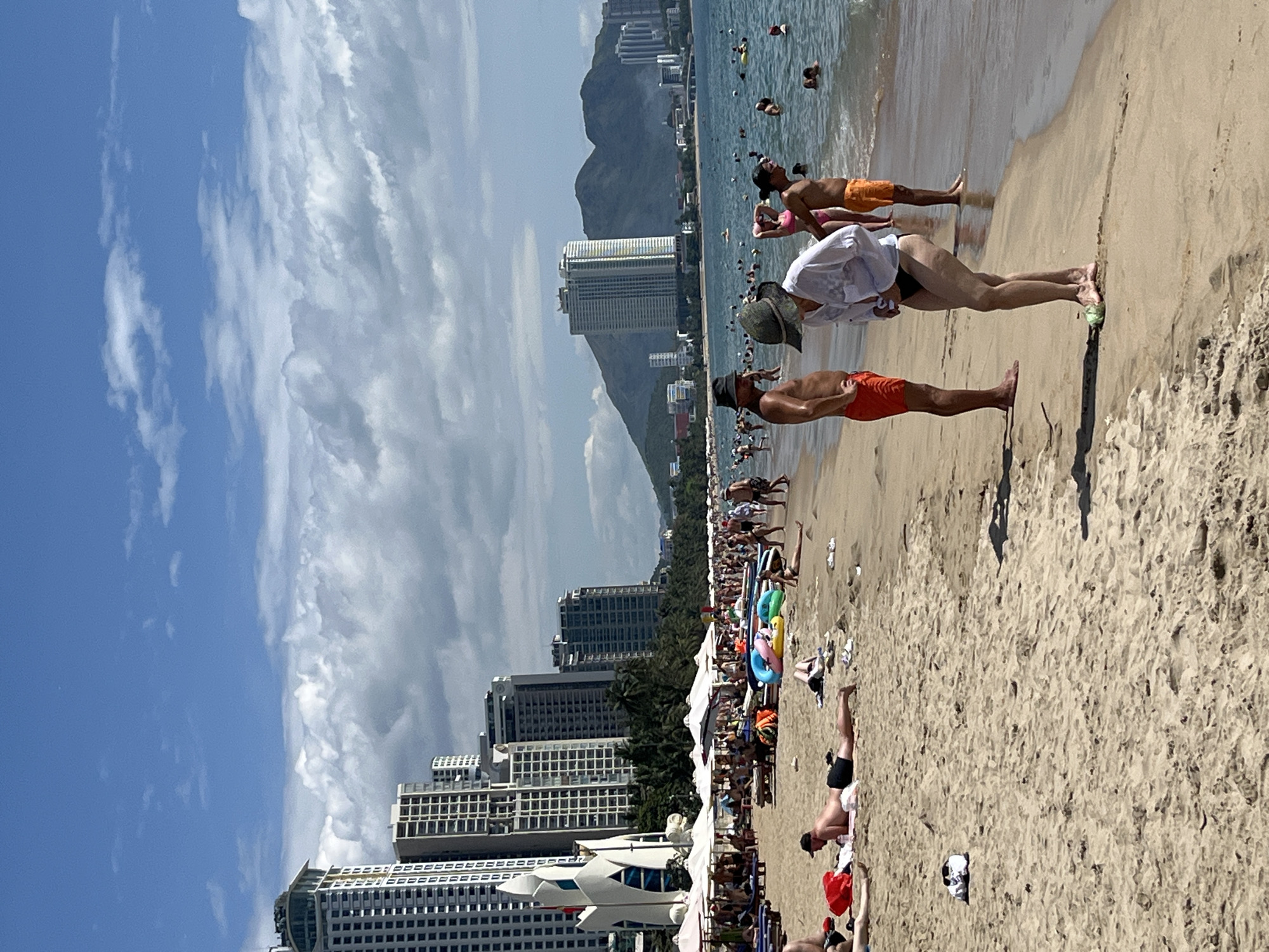 This image depicts a bustling beach scene in Nha Trang, Vietnam, characterized by its sandy shore, the clear blue water of the ocean, and a backdrop of modern high-rise buildings. The beach is populated with numerous people engaged in various activities: some are sunbathing on lounge chairs and towels, others are strolling along the shoreline, and many are swimming or wading in the ocean. In the background, a series of tall, contemporary buildings line the coast, reflecting the urban development of Nha Trang. Scattered on the sand are beach umbrellas, inflatable toys, and personal belongings, contributing to the lively atmosphere. The sky is a mix of blue and white clouds, suggesting a pleasant, sunny day. The overall impression is one of a popular tourist destination, where both natural beauty and urban infrastructure coexist.