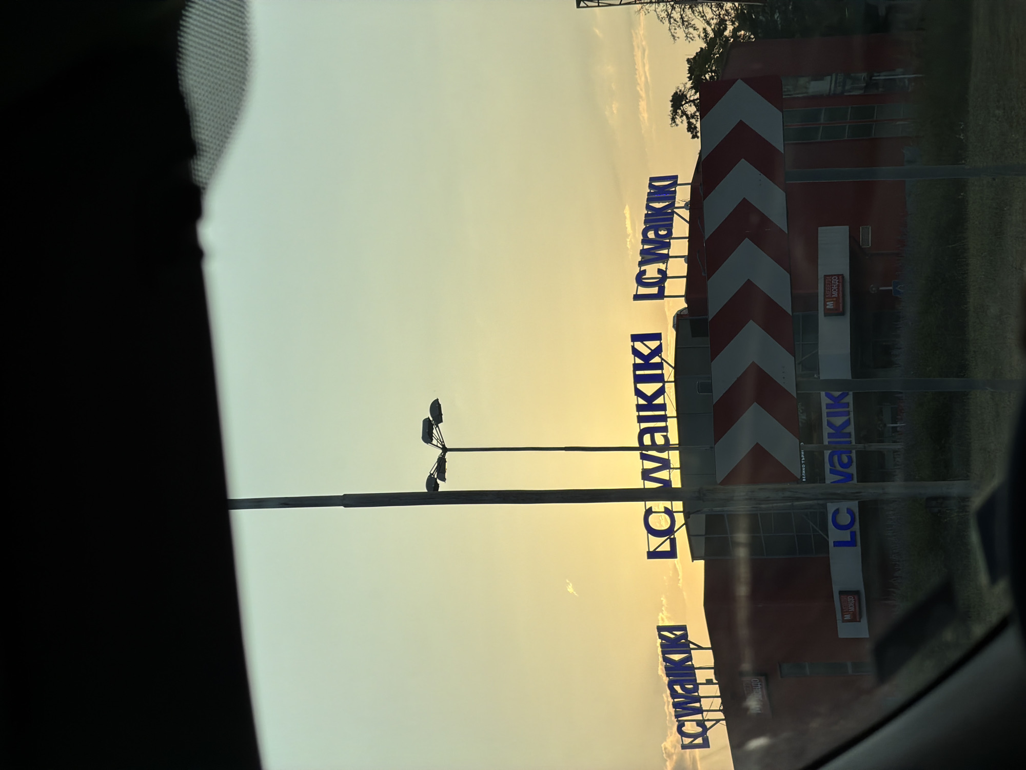 The image shows a reflection of a building and signage in what appears to be a vehicle's windshield. The building is red with white and red chevron markings on the side, suggesting it is a commercial establishment. Large blue letters spelling "LC Waikiki" are visible, indicating a clothing store. There are also smaller signs on the building, one of which says "M MONDO" in red and white. A street light with three lamps is also present. The sky is a pale yellow, suggesting either sunrise or sunset. The overall scene is illuminated by warm, soft light. The text "LC Waikiki" is repeated multiple times, vertically oriented due to the reflection. The location context provided is Veliko Tarnovo, Bulgaria. The image is a photograph taken from inside a vehicle, as evidenced by the dark frame on the left and the curved line of the windshield.