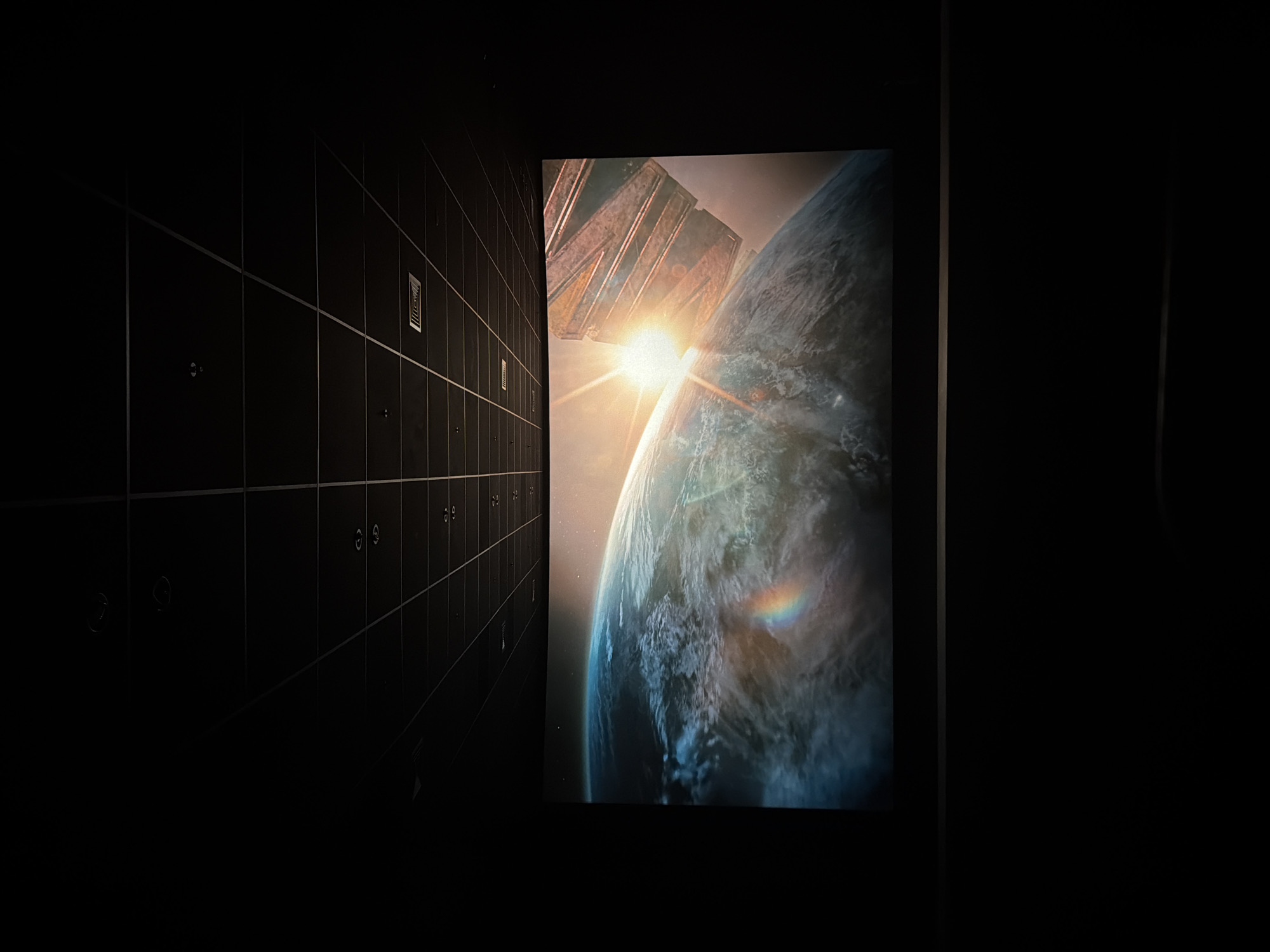 The image shows a large cinema screen displaying a scene of a planet, possibly Earth, with a bright sunburst and a large, metallic structure in the foreground. The screen is curved and occupies the majority of the lower half of the frame. Above the screen, a dark, tiled ceiling with recessed lighting and vents is visible. The overall environment is dark, typical of a movie theater. The image appears to have been taken from a seating area within the theater, as suggested by the dark foreground which could be seats or the floor. The scene on the screen is a realistic rendering, possibly from a science fiction film or documentary. There are no people visible in the image. The location is not specified in the image itself and cannot be determined without additional information.