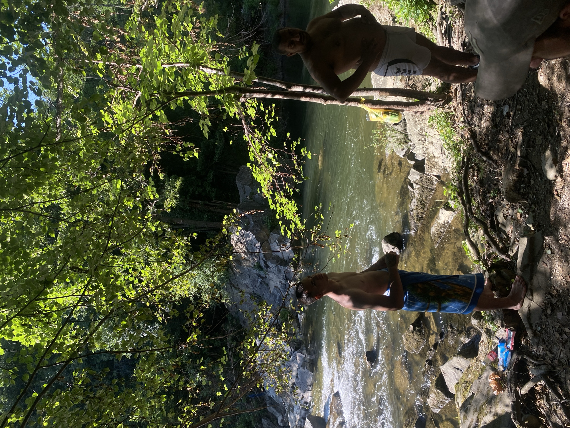 The image depicts an outdoor scene near a river, possibly in Debnevo, Bulgaria, given the context. The foreground is dominated by a rocky, wooded area with dappled sunlight filtering through the leaves of trees. Two shirtless men are present. The man on the left is younger, wearing a blue towel wrapped around his waist, and is looking upwards. He is standing on rocks near the river. The man on the right is older, wearing white athletic shorts, and has his hands on his hips. He is standing on the shaded bank. A yellow plastic bag containing what appear to be green items is hanging from a tree behind him. A river with flowing water and rocks is visible in the background, with a lush, green forest and rocky cliffs beyond. On the ground near the man on the left, there are several discarded items including a red bottle and wrappers. A person's head wearing a baseball cap is visible in the extreme lower right corner of the image, suggesting a fourth individual is present but mostly out of frame.