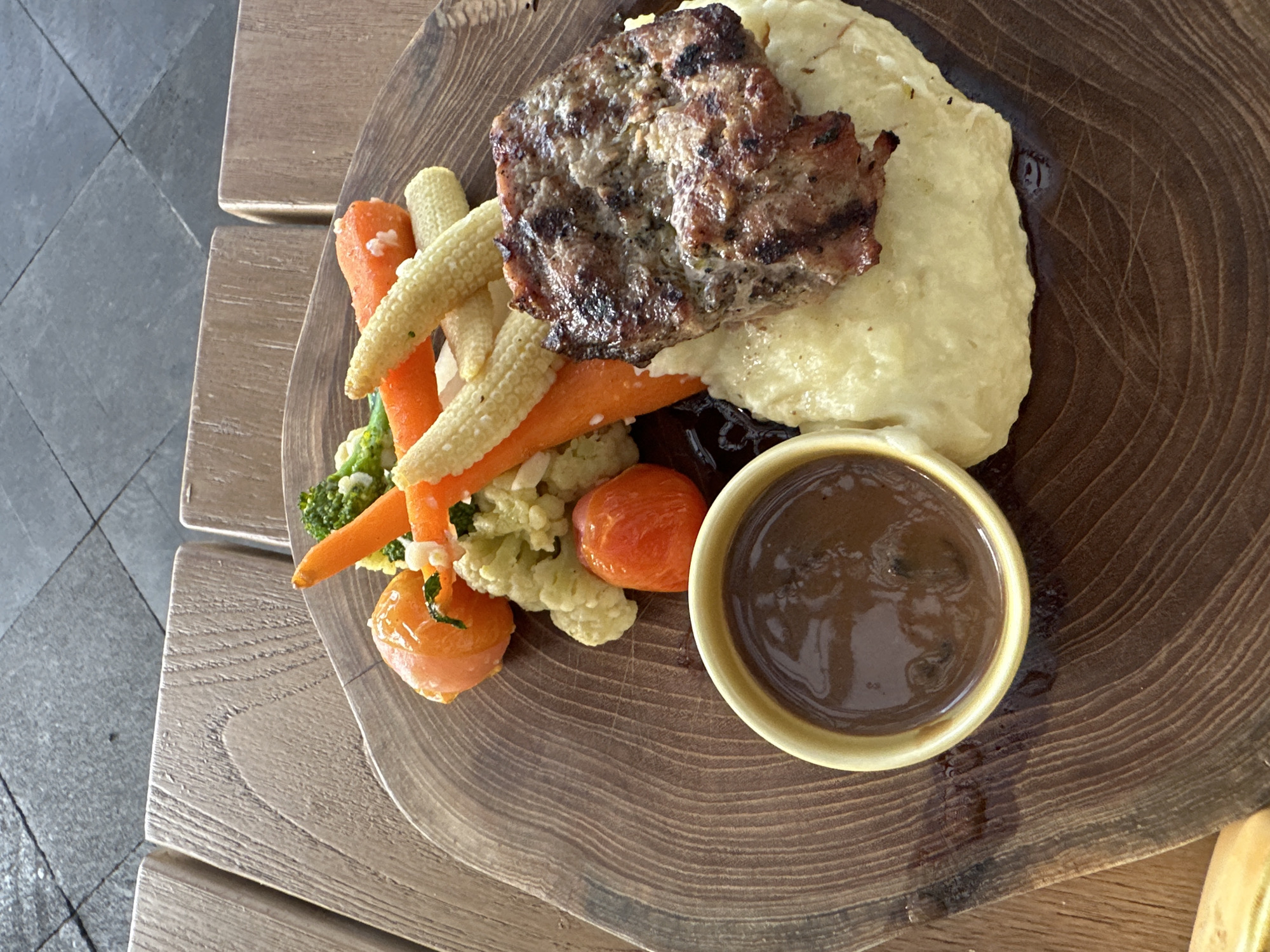 A close-up, top-down shot shows a meal served on a rustic, round wooden plate. The plate rests on a slatted wooden surface, with a darker tiled surface visible in the background. The meal consists of mashed potatoes, a grilled meat patty, a side of mixed vegetables including carrots, corn on the cob, cauliflower, and broccoli, and a small bowl of dark sauce. The lighting suggests it is daytime, possibly outdoors given the natural materials and the texture of the tiling, which could be a patio or outdoor dining area in Kuta Selatan, Indonesia. There are no people visible in the image.