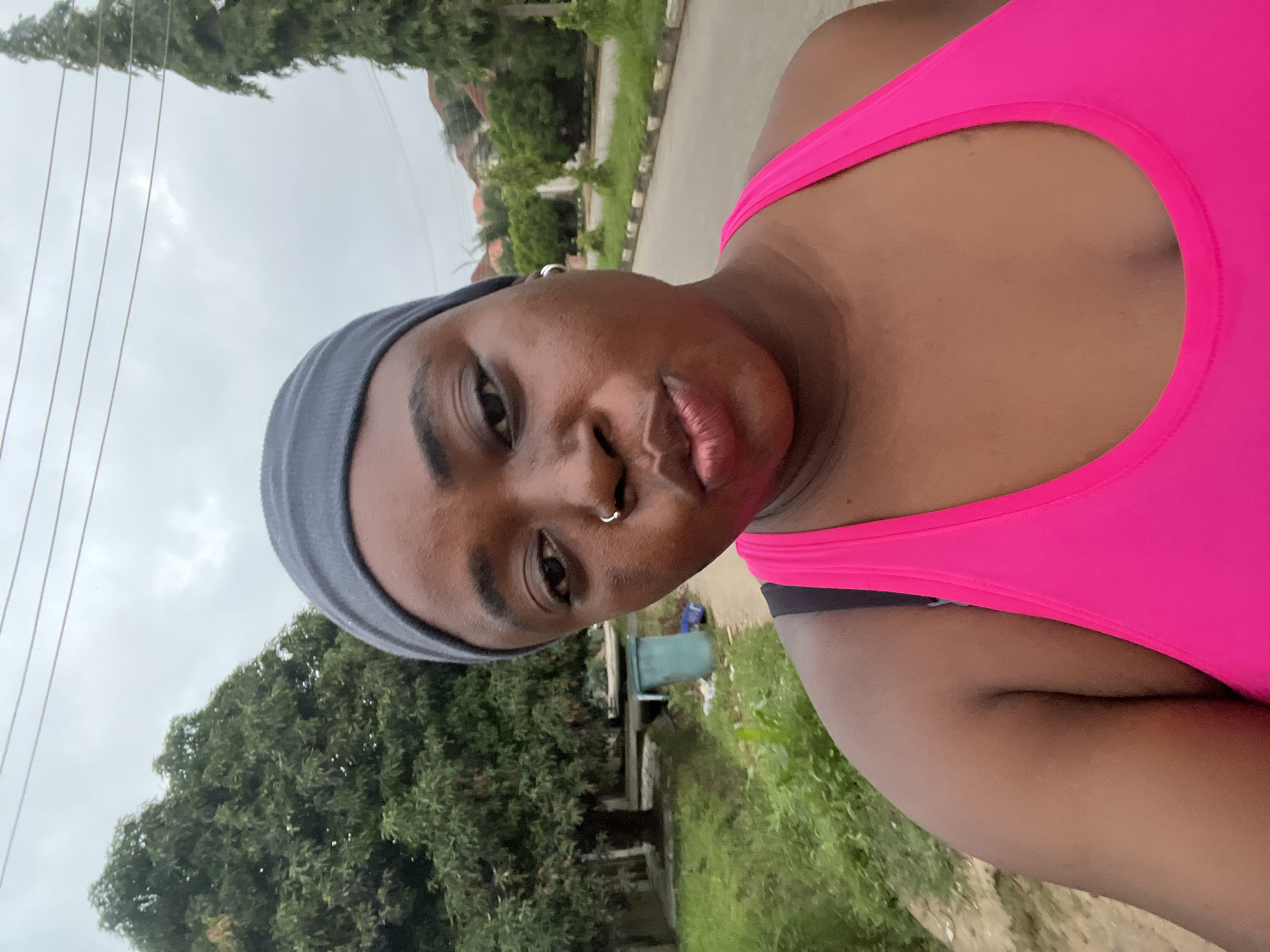 An outdoor, medium shot, selfie-style image shows a young Black woman in a bright pink athletic top and a grey headband. She has a silver hoop earring in her left ear and a silver nose ring. The woman is looking directly at the camera with her lips puckered. Behind her, a tree-lined residential street is visible under an overcast sky. The street has a paved road and a sidewalk with grass and some bushes. A green trash can is visible on the left side of the image, near a ditch. The environment appears to be a residential area within the city of Abuja, Nigeria.