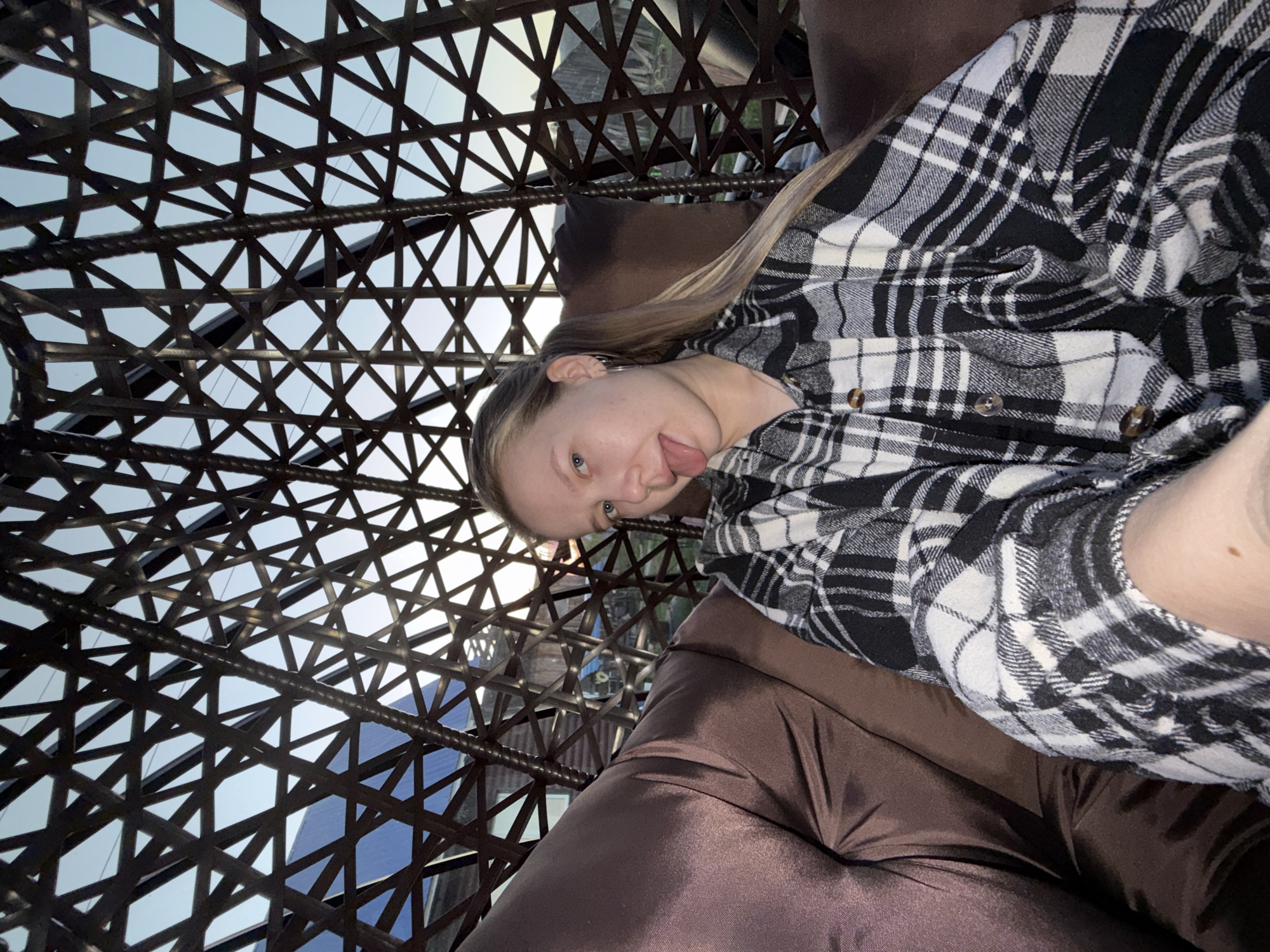 A young woman is sitting in a wicker chair. She is wearing a black and white flannel shirt and sticking her tongue out. The chair is in an outdoor location. The sky is visible through the wicker pattern of the chair. Based on the location requested, the scene is purported to be in Zhigalki, Russia.
