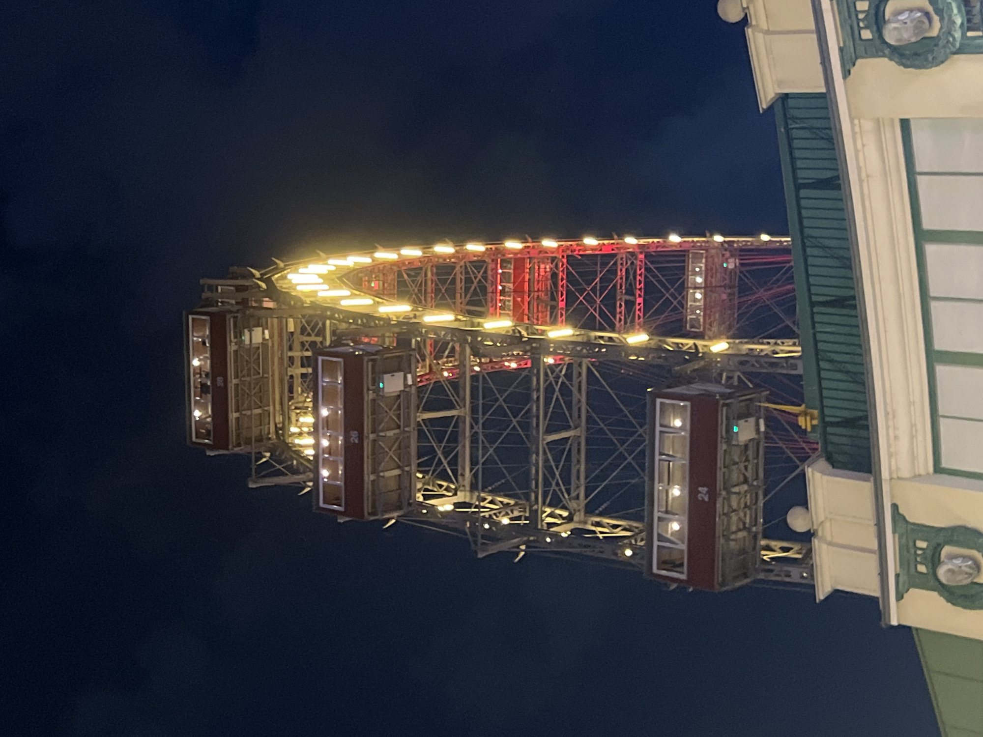 The image shows a close-up, low-angle nighttime view of the Riesenrad, a Ferris wheel located in Vienna, Austria, specifically in the Prater amusement park. The iconic Ferris wheel is illuminated with yellow and red lights, with several of its passenger cabins visible. One cabin in the foreground displays the number "24". The dark night sky provides a backdrop to the illuminated structure. In the lower portion of the image, the roof and facade of a building, possibly part of the amusement park or an adjacent structure, are visible. This building has decorative elements, including what appears to be a wreath with a relief sculpture.
