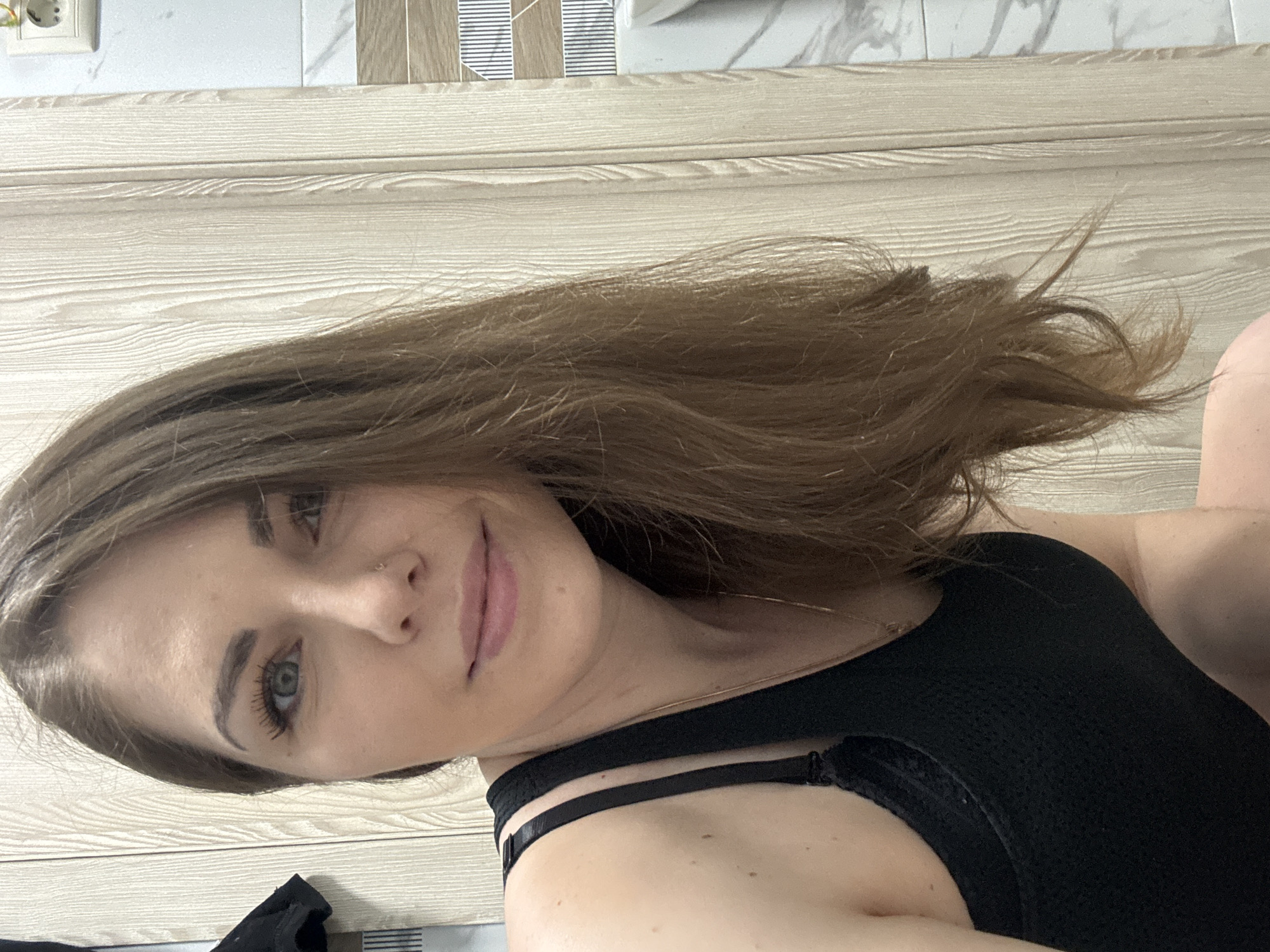 The image is a selfie of a young woman with fair skin, light brown hair, and blue eyes. She is wearing a black top with thin straps. The background consists of a wall with light-colored wood paneling and a section of tiled wall near the top with a power outlet visible in the upper left corner. The setting does not provide any evidence of the location being in the city of Раздолье, Russia.