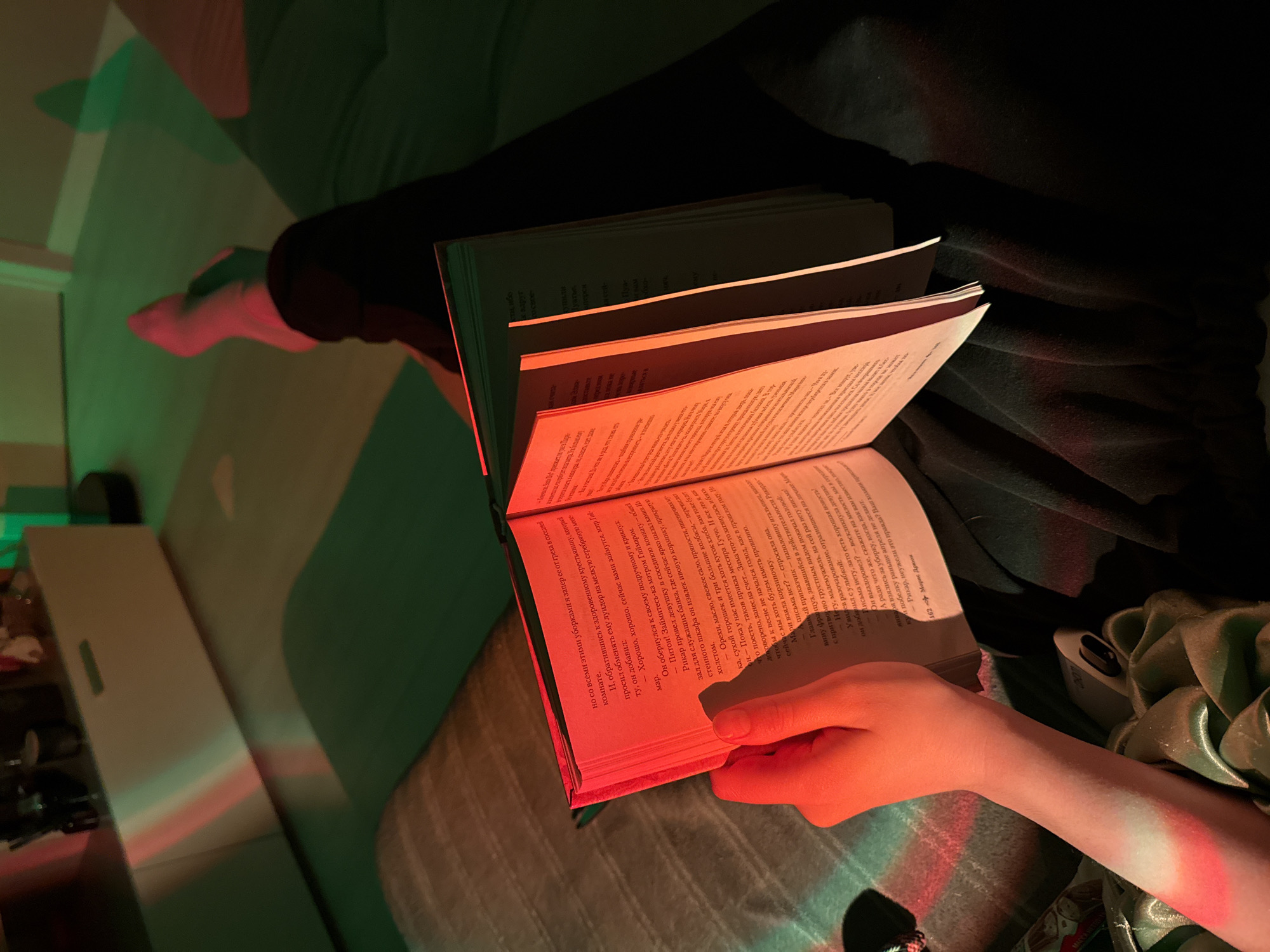 A person is sitting in an unknown location, with legs outstretched on a green surface. The person is holding an open book, with red light illuminating the pages. The hand is holding the book open. The room is illuminated with green and red lighting, creating an unusual atmosphere. A white cabinet is visible in the background.
