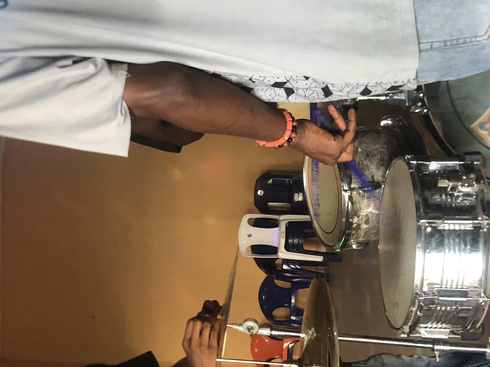 A person is playing a drum set. The drum set includes a snare drum and a cymbal. There are stacked plastic chairs nearby. The location is Nkwelle Ezunaka, Nigeria.