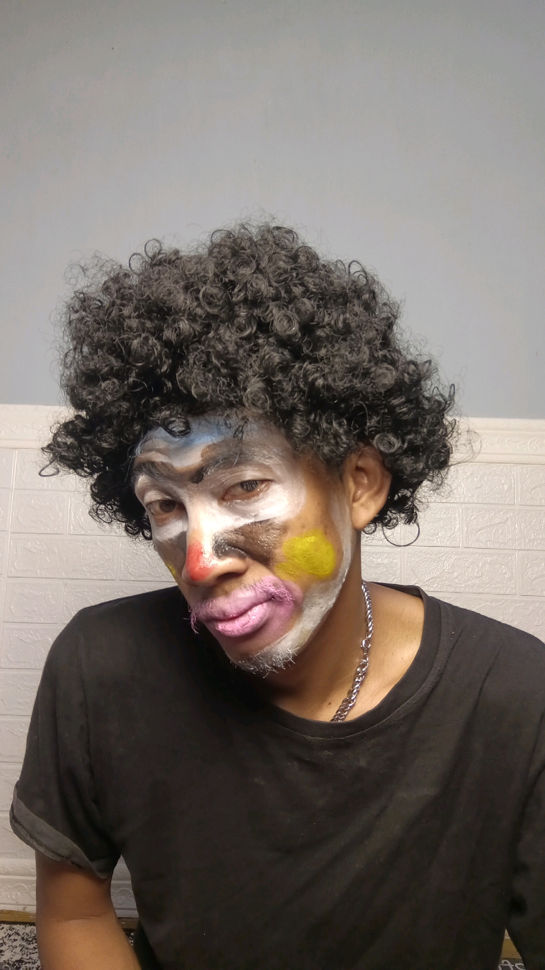 A person with black, curly hair is wearing clown makeup, including white on their face, red on their nose, yellow on their cheek, and pink on their lips. They are wearing a black t-shirt and a silver chain necklace. The background consists of a light-colored tiled wall and a plain wall. The person appears to be indoors, possibly in a domestic setting within Kebumen, Indonesia.