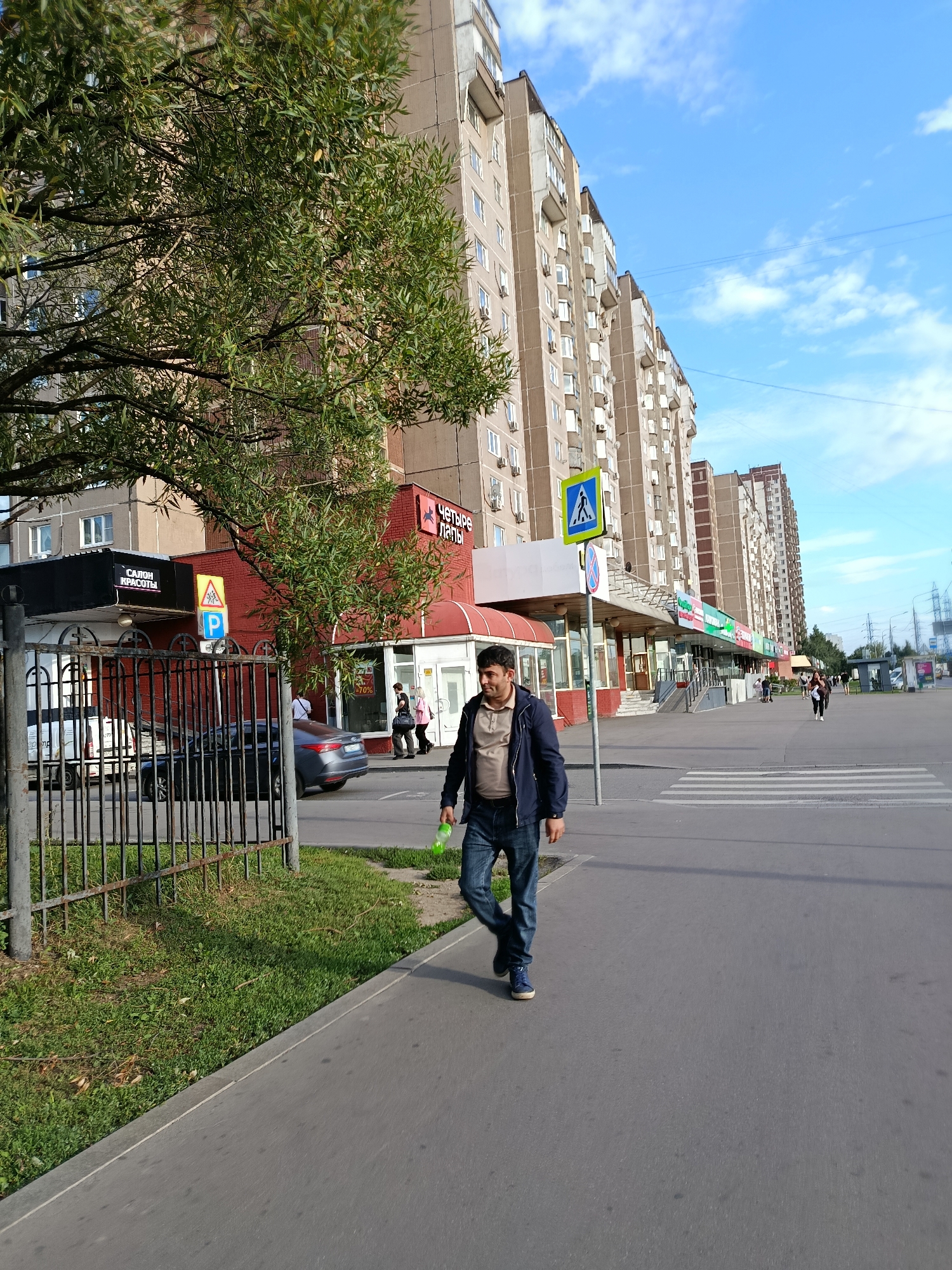 A medium-wide shot captures a man walking on a sidewalk in front of a row of apartment buildings in Moscow, Russia, on a sunny day. The man, wearing a dark blue jacket over a tan shirt and blue jeans, is walking towards the right of the frame, holding a green plastic bottle in his right hand. He appears to be in his late 30s or early 40s with dark hair and a mustache.
The apartment buildings are tall and light brown, with numerous windows and air conditioning units visible. The ground floor of the buildings features a series of shops and businesses. One shop has a red facade with Cyrillic lettering that translates to "Four Paws." Another sign above a black storefront reads "Салон Красоты," meaning "Beauty Salon." A parking sign is visible near the beauty salon.
A pedestrian crossing is marked by white stripes on the asphalt road in front of the shops. A metal fence borders the sidewalk on the left side of the image, separating it from a grassy area. A portion of a dark-colored car is visible behind the fence, parked on the street. The sky is blue with scattered white clouds, and overhead power lines are present in the distance. The overall scene depicts a typical urban environment in Moscow.
