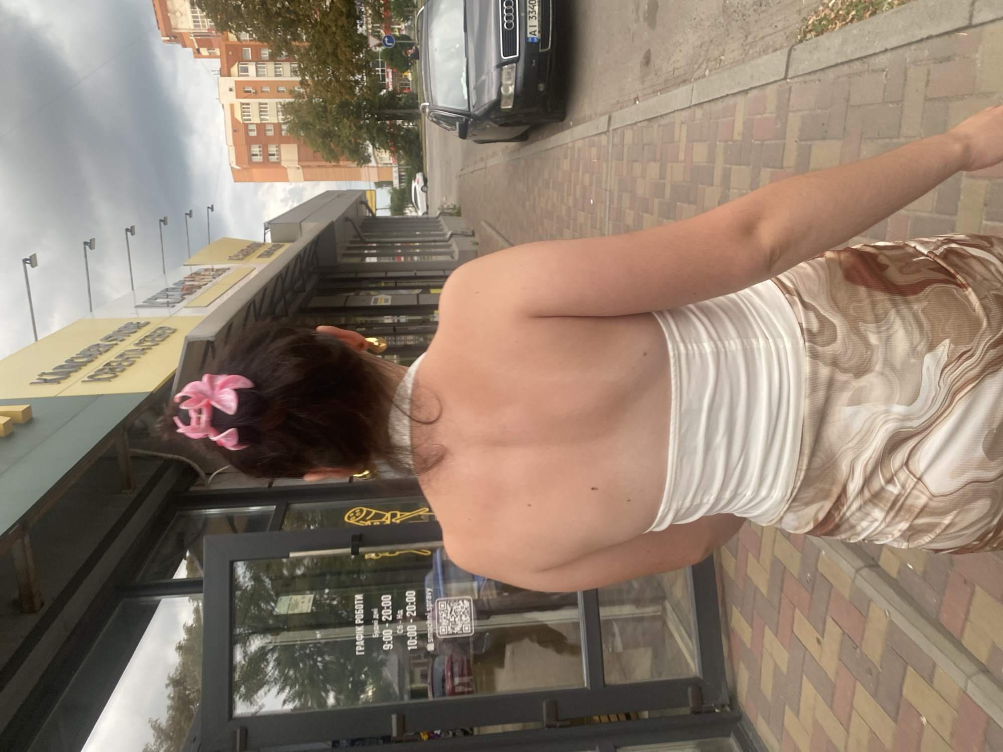 The image depicts a woman from the back, standing on a sidewalk in Poltava, Ukraine. She has dark hair pulled up with a pink bow and is wearing a white halter top and a brown and white marble-patterned skirt. She is facing away from the camera and towards a building with a storefront. The storefront has a glass door and signage in Cyrillic script that appears to indicate business hours. To the right of the woman, a black Audi sedan is parked on the street. In the background, there are buildings, trees, and a cloudy sky. The sidewalk is paved with brick-like stones.