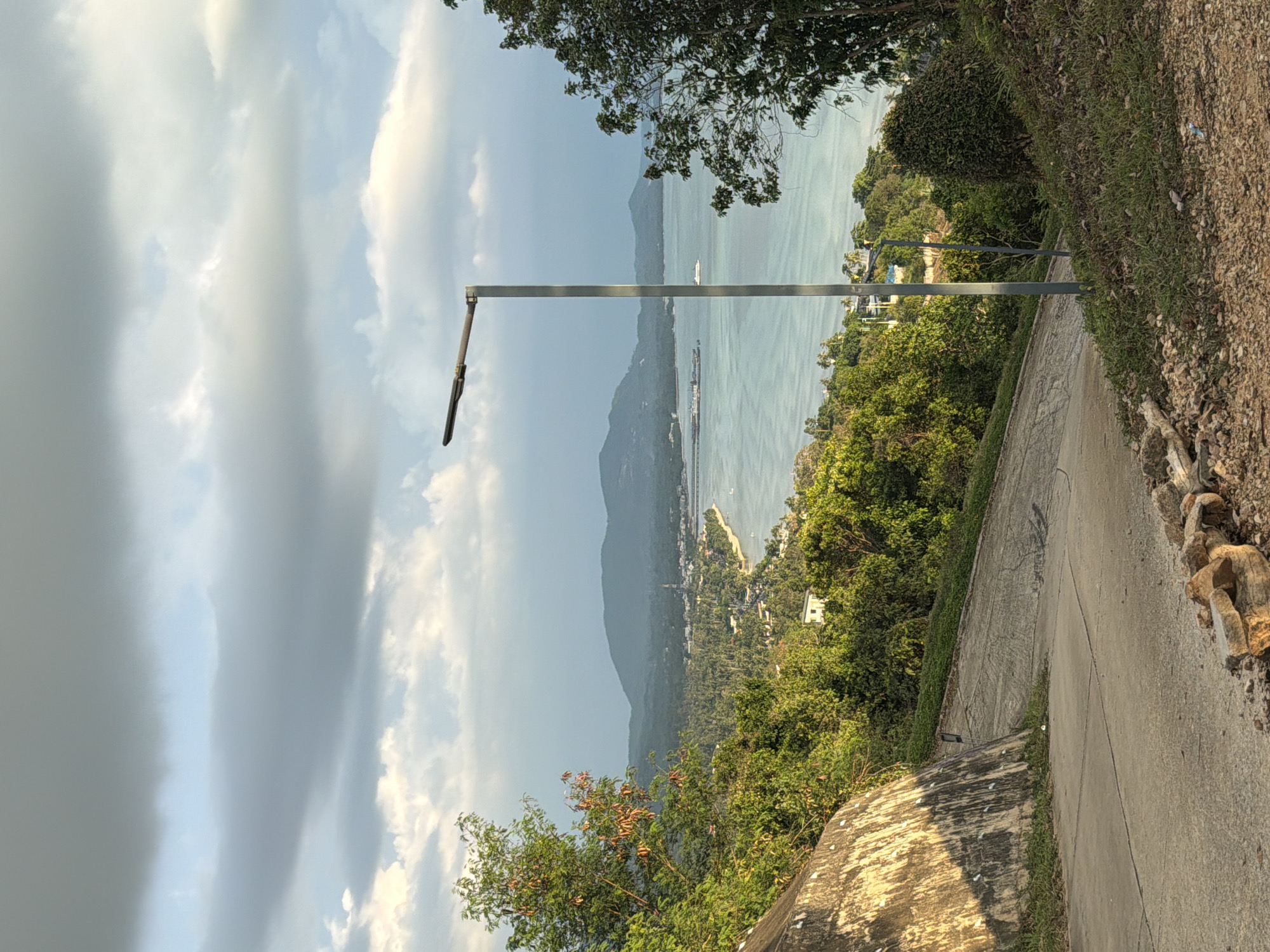 The image depicts a scenic overlook in Baan Thong Po, Thailand, featuring a winding concrete road leading down towards a coastal town nestled between lush green hills and the turquoise ocean. A modern, silver lamppost stands prominently on the right side of the road, its single light fixture extending outwards. The sky above is a mixture of light blue and dramatic grey clouds, hinting at changing weather. In the distance, mountains form a soft, hazy backdrop to the bustling town and the calm sea. Several boats are visible on the water, and the coastline is dotted with buildings and dense vegetation. The foreground of the image is dominated by the road, a rough-hewn stone wall on the left, and a patch of dry grass and gravel on the right.