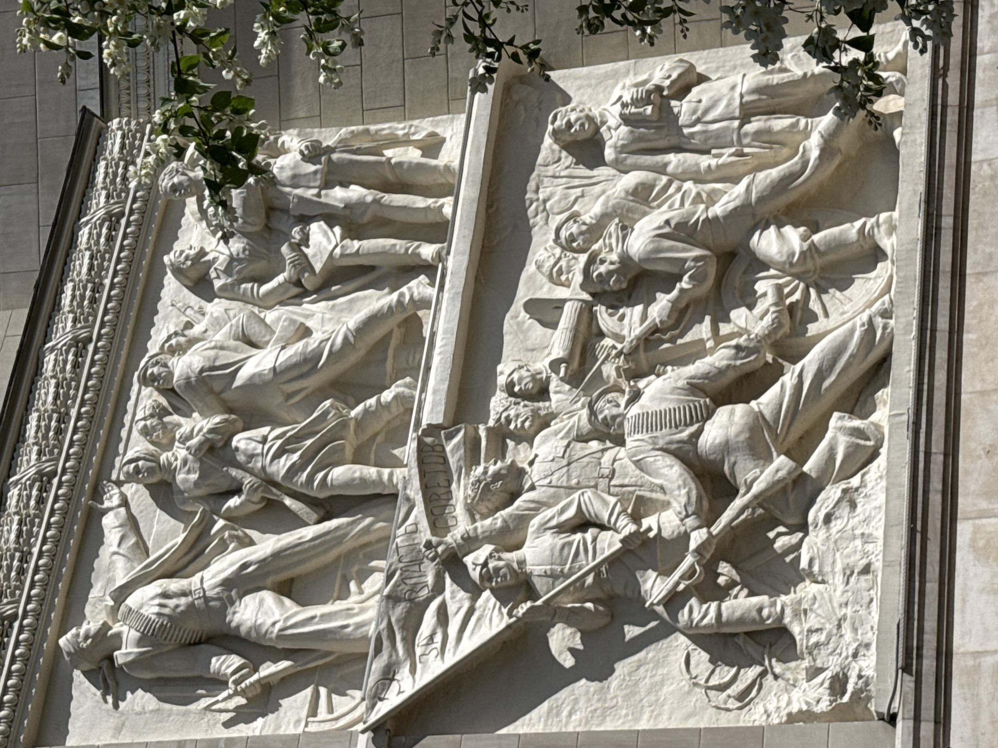 The image depicts a dynamic, white bas-relief sculpture adorning the exterior of a building. The sculpture features a chaotic scene of many figures, appearing to be soldiers or revolutionaries, in various states of action and distress. They are depicted in relief, with figures overlapping and tumbling over one another, suggesting a moment of intense struggle or conflict. Some figures are shown lying down, some are reaching out, and others are holding weapons like rifles or sabers. The clothing suggests a historical period, possibly early 20th century.
The overall impression is one of struggle, perhaps a battle or an uprising. The figures' expressions, though difficult to discern in detail due to the relief format and distance, seem to convey urgency and effort. The lighting suggests it is daytime, with strong shadows casting depth and drama onto the sculpted forms. There are some white flowers and green leaves hanging from the top of the frame, possibly from a tree or a decorative element on the building, which contrasts with the starkness of the sculpture. The building's facade, made of pale stone blocks, provides a neutral backdrop to the detailed relief.
The sculpture is divided into two main panels, separated by a vertical element. On the left panel, figures are arranged in layers, some appearing to fall or be overcome. The right panel shows a similar density of figures, with a prominent banner being held aloft by one individual, and another figure gripping a rifle. Visible text on the banner is in Cyrillic script, with partial words like "СОВЕТОВ" (SOVETOV, likely part of a longer phrase related to Soviets) and "ЗА" (ZA, meaning "for"). This text, along with the historical attire and the representation of struggle, strongly suggests a Soviet-era monument, likely commemorating a revolutionary event or a significant battle. Given the location context of Moscow, Russia, this is a monument of historical and ideological importance.