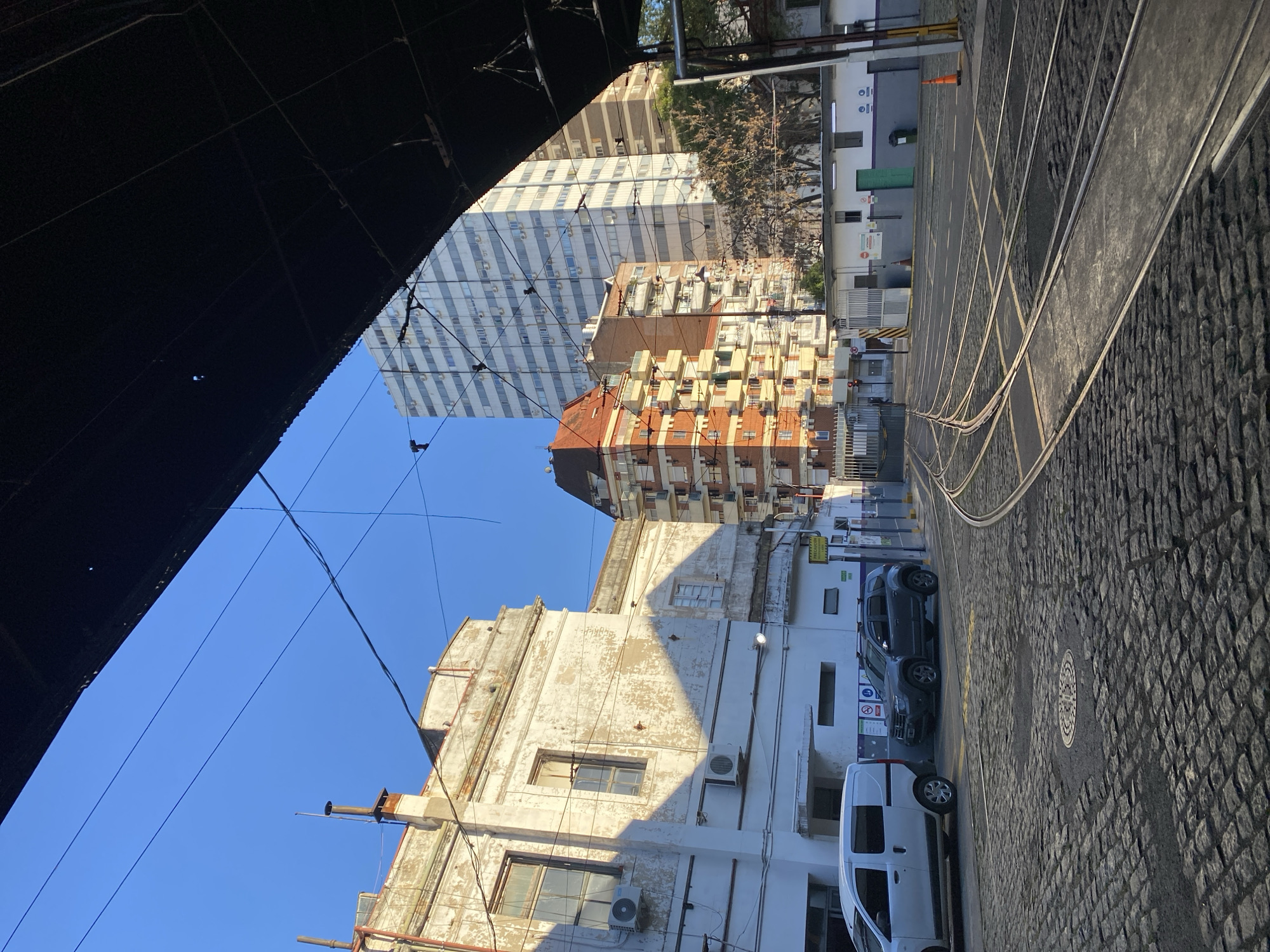 The image, taken at an extreme angle, depicts a street scene in Buenos Aires, Argentina. The perspective is from beneath an overhead structure, possibly a bridge or elevated platform, which obscures the upper left portion of the frame.
The street, paved with cobblestones, runs diagonally from the lower right to the upper left, its perspective converging into the distance. Tram tracks are embedded in the cobblestones, indicating a streetcar line. Vehicles are parked along the right side of the street, including a white van and a gray SUV.
Several buildings of varying styles and heights line the street. A tall, white apartment building with numerous windows is visible in the background. Closer to the camera is an older, weathered building with a peeling facade. A building with red brick and numerous balconies can be seen between the apartment building and the peeling facade.
Overhead power lines crisscross the sky. The sky is clear and blue. The strong shadows cast on the buildings suggest a sunny day. The city of Buenos Aires is the location, based on the architectural styles and street features visible.