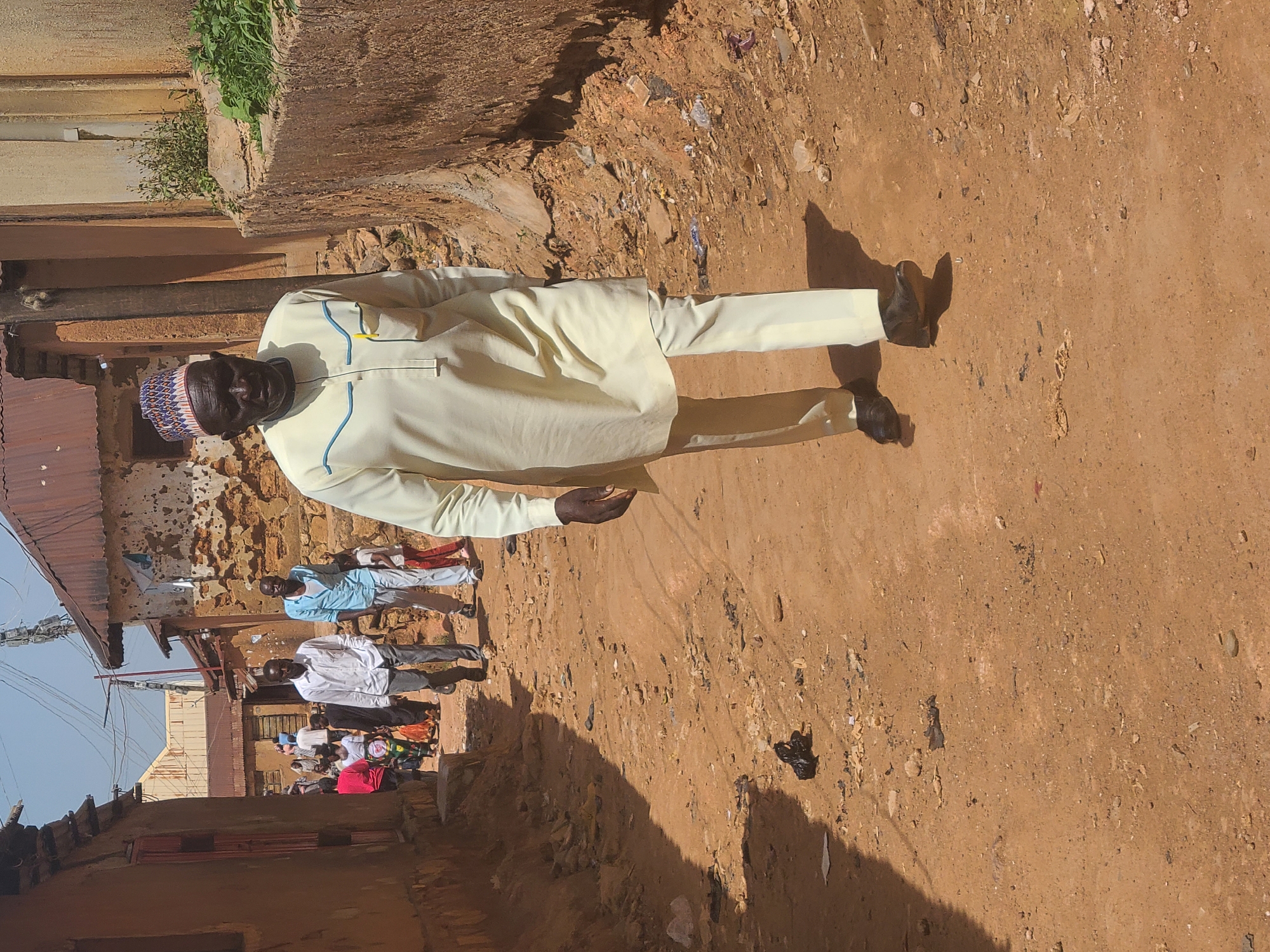 A full-length photograph shows an older man standing on a dirt road. The man is wearing a light yellow outfit with blue trim and a traditional cap. Shadows suggest the photo was taken in bright sunlight. The ground is uneven and littered with debris. In the background, other people are visible, along with buildings and structures that appear to be old or in disrepair. The location appears to be a street in a densely populated area.