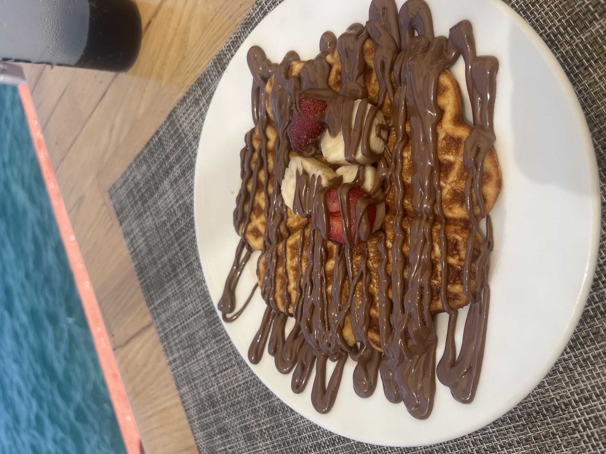A plate of waffles with chocolate syrup and fruit is on a table with a view of the sea. The waffles are arranged in a circular pattern on a white plate. The plate is drizzled with chocolate syrup and topped with sliced bananas and strawberries. In the background, the turquoise water of the sea is visible, with a railing and a wooden table. A glass of dark liquid, possibly soda, is on the right side of the table. The scene suggests an outdoor dining experience in Alanya, Turkey, likely at a restaurant or cafe with a seaside location.