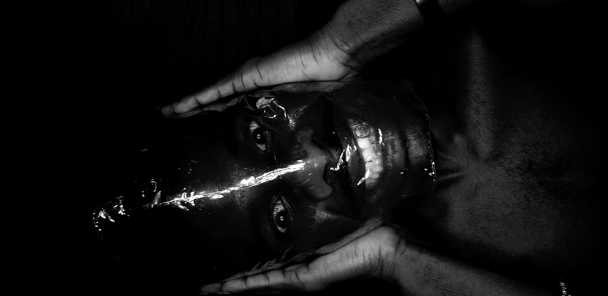 This black and white, high-contrast photograph presents a close-up of a person's face largely obscured by crinkled, reflective plastic material, possibly a clear plastic bag or packaging. Two dark-skinned hands frame the face, one at the top and one at the bottom, pressing or holding the plastic against it. The person's eyes are visible through the transparent material, wide open and looking directly forward, exhibiting a strong, intense gaze.
The activity depicted appears to be a staged portrait or conceptual art piece, exploring themes of suffocation, identity, or concealment. The dark background isolates the subject, emphasizing the struggle or containment suggested by the plastic. Bright, linear reflections on the crinkled plastic highlight its texture and the contours of the face beneath. The monochromatic palette intensifies the dramatic and somber mood of the image.
Visible text on the plastic is faint and distorted due to the crinkles and reflections. In the upper right quadrant, a circular logo appears to contain "200R" or "200P" and possibly the word "RED," alongside traces of "recycle" or "recyclable." Along a prominent horizontal reflection line across the face, fragmented words like "Salty" or "Salad" are partially discernible, suggesting the material could be from food packaging. Other scattered, illegible text and numbers appear across the surface. The scene is devoid of specific location cues beyond the implied setting of Narayi, Nigeria through the subject's appearance.