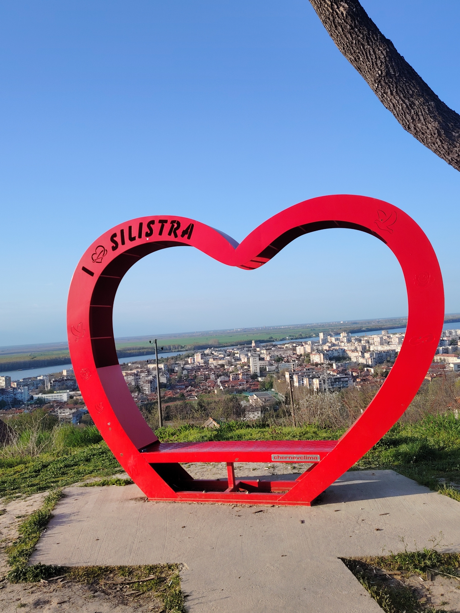 A view of the city of Silistra is seen through a large red heart-shaped frame. The frame has the words "I ❤️ SILISTRA" at the top. The structure also serves as a bench seat. A body of water and green fields can be seen in the distance, beyond the city. A tree branch is visible in the upper right corner of the frame. The bench rests on a concrete base, surrounded by grass and dirt. The sky is a clear blue.