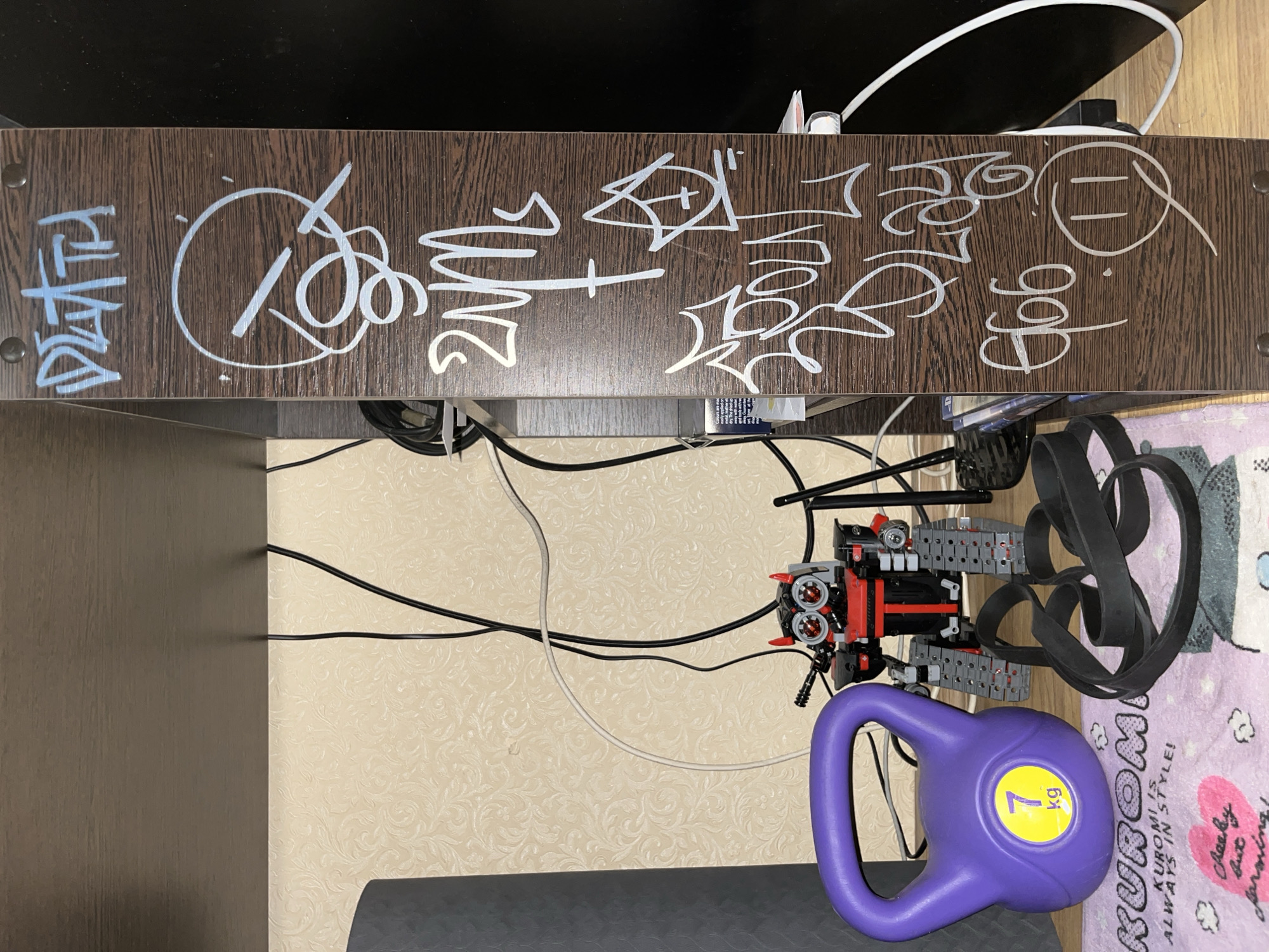 The image shows a close-up of a dark brown wooden shelf with graffiti on its side. The graffiti is done in white and light blue paint and appears to be tags or stylized lettering. Some of the visible text includes "DEATH," a circular design with lines and loops, "2NM," a symbol with a cross inside a triangle, and "SOUL PLAY." At the bottom, there is "€66" and a smiley face drawing.
To the left of the shelf, partially visible objects include a purple kettlebell with a yellow "7 kg" sticker, a black resistance band, and a pink towel with cartoon characters and text that says "KUROMI IS ALWAYS IN STYLE!". Behind these objects, a toy robot with red and black details is visible. Various black and white cables are also present, running across the background and connecting to unseen devices. The wall in the background has a light-colored patterned wallpaper. The overall setting appears to be an indoor space, possibly a room or a storage area. There are no people visible in the image.