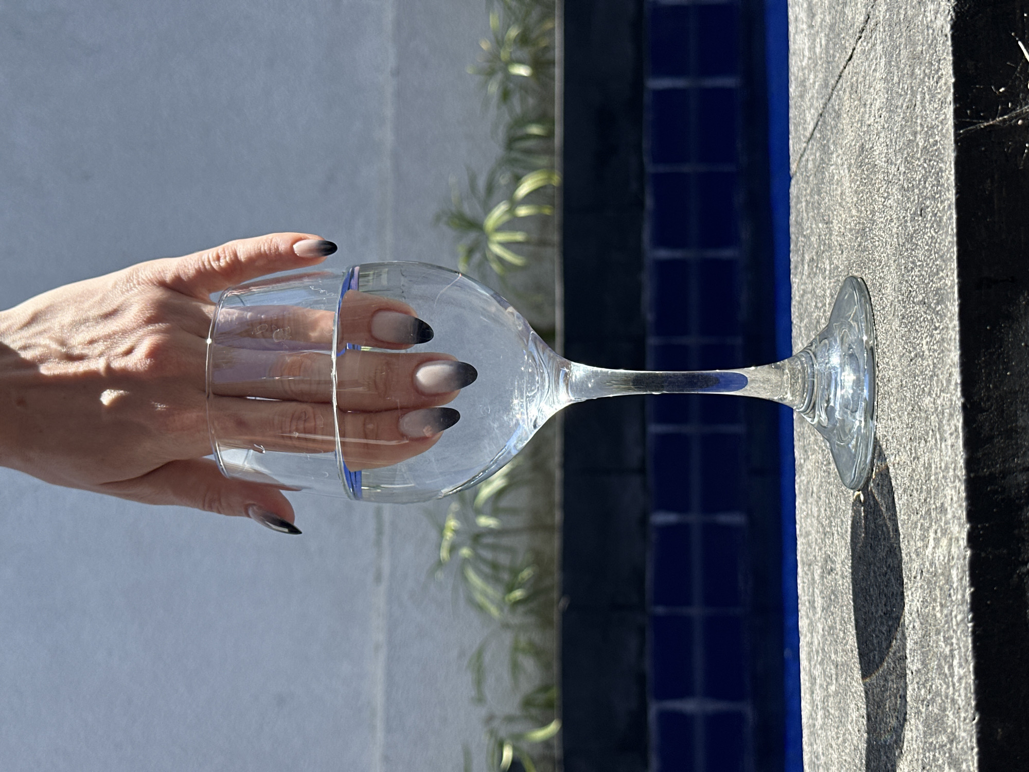 A close-up, eye-level shot features a person's hand with ombre black and grey nail polish dipping into a clear glass of water. The glass is positioned on a textured gray surface, casting a distinct shadow. In the background, a blue tiled swimming pool is visible, with green foliage and a light blue wall beyond. The scene suggests an outdoor setting, possibly in North Kuta, Indonesia.