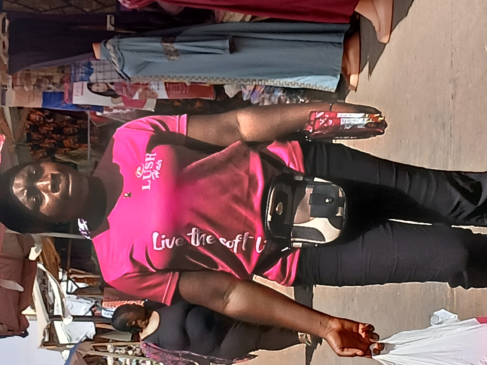 This image, taken in Maiburiji, Nigeria, captures an outdoor market scene during the daytime under bright, clear skies. The primary subject is a person of color, standing with a neutral expression while looking towards the camera. They are wearing a vibrant pink t-shirt, dark pants, and a black and camouflage fanny pack around their waist.
The pink t-shirt prominently features white text: "LUSH Hair" with a crown logo above it, and below that, "Live the soft li" (likely part of "Live the soft life"). In their right hand, the person holds a red and silver packaged item, identifiable as a can of Pepsi. Their left hand holds a crumpled white plastic bag.
In the background, another person in a dark top is partially visible. The overall setting reveals a bustling market environment with numerous stalls displaying clothes and other merchandise hanging on racks and lines. The ground beneath appears to be a paved or concrete surface.