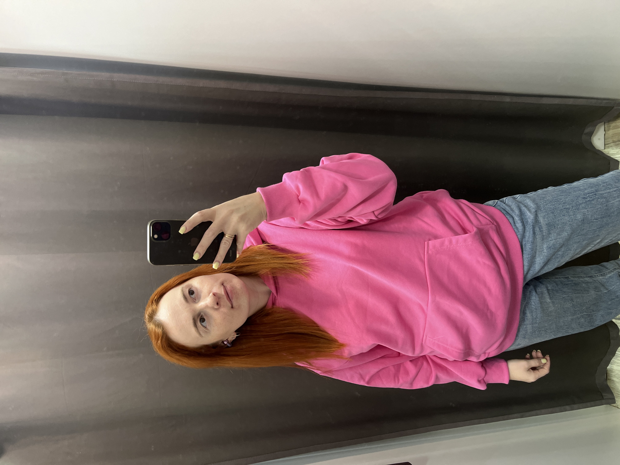 The image shows a young woman with long, auburn hair taking a selfie in what appears to be a fitting room or a room with curtains. She is wearing a bright pink oversized hoodie with a front pocket and light blue denim jeans. Her left hand is holding a black smartphone, and her right hand is resting on her hip, displaying a yellow nail polish. She has a subtle expression of contemplation on her face. The background consists of dark gray curtains to her left and a white wall to her right. The flooring appears to be light-colored wood. The overall lighting suggests an indoor setting, likely a retail store. There is no visible indication of the city of Chaikovsky, Russia, or any other specific geographical location.
