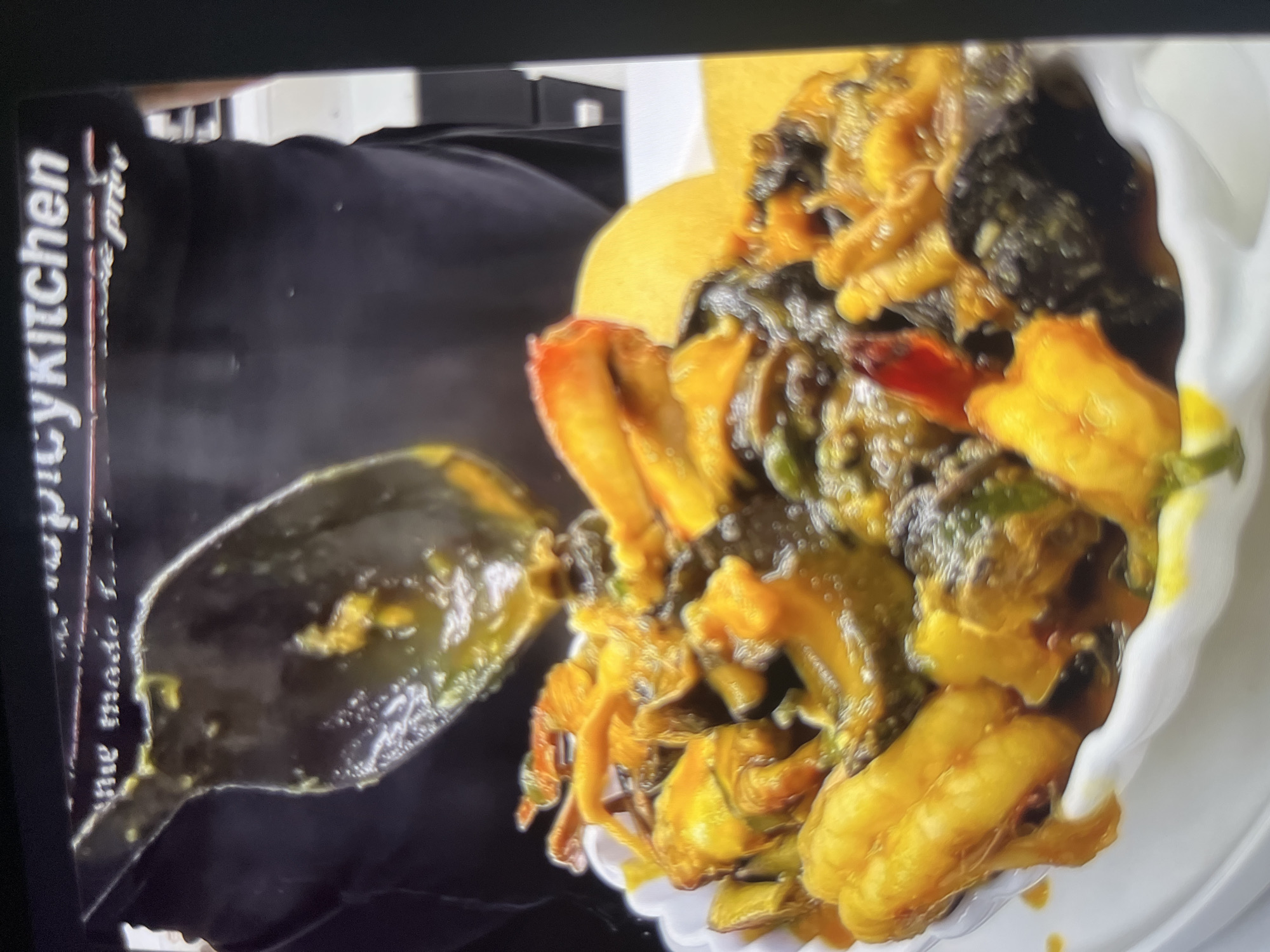 The image shows a close-up of a white bowl filled with a colorful, saucy dish. The dish appears to be a stew or curry containing pieces of seafood, possibly shrimp and crab, along with some vegetables. The sauce is a vibrant yellow-orange color. In the background, a person wearing a black t-shirt with white text that reads "spicykitchen" is visible. The person is holding a large ladle over the bowl, suggesting they are serving or presenting the food. The overall impression is one of a delicious, home-cooked meal. There is no information in the image that provides any context for the city of Abuja, Nigeria.