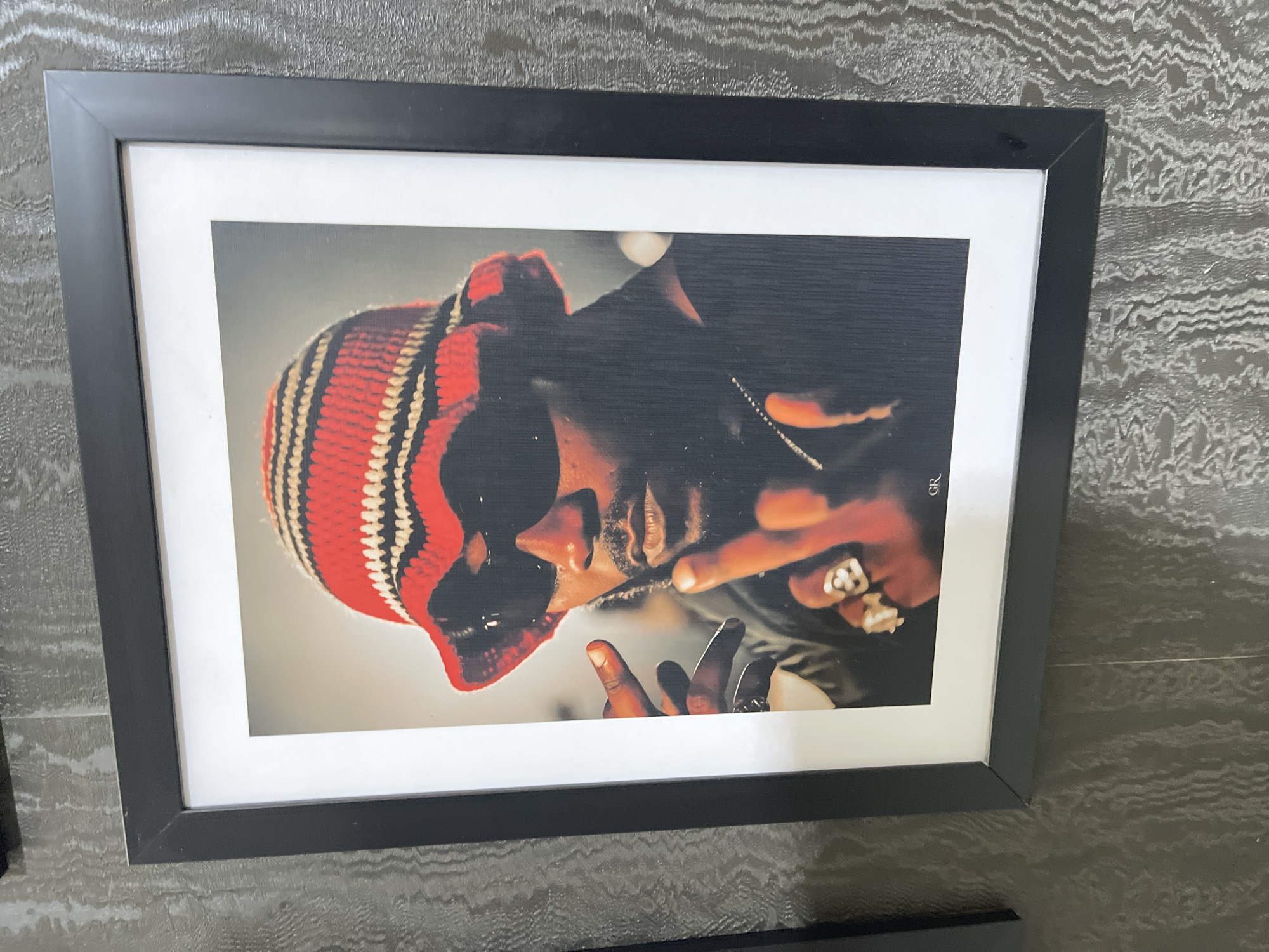 A photograph captures a black-framed picture hanging on a textured, dark grey wall. The picture, which is slightly rotated, prominently displays a portrait of a person from the chest up. The person has dark skin and a visible beard, and wears a distinctive knit hat (possibly a beanie or balaclava) striped in red, black, and off-white, covering part of their forehead and ears. Multiple rings adorn their fingers, and a chain necklace is visible. The person's right hand is raised near their face, with two fingers extended in a gesture. The lighting in the portrait is dramatic, creating deep shadows and highlighting features. The background within the portrait is a soft gradient from light to dark grey. In the top right corner of the print, just inside the white matting, the letters "GR" are visible. The black frame encloses the print, which is set off by a wide white mat. The wall on which the picture hangs features an intricate, wavy or fibrous pattern in various dark grey tones, giving it a rich, textural appearance. A subtle reflection can be seen on the frame's glass. Part of another dark, possibly framed object is visible at the bottom edge of the overall image.
