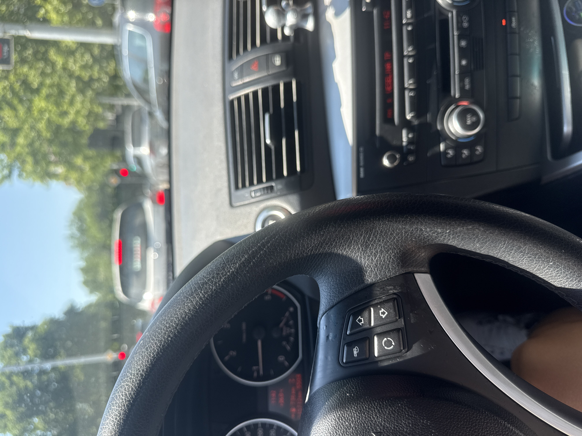 The image depicts the interior of a car, likely a BMW, with a driver's perspective on a sunny day. The steering wheel is visible on the left, with control buttons. The dashboard displays a speedometer and a central console with air vents and a car stereo. Outside the car, traffic can be seen, with several other vehicles on the road. The scene suggests the car is in traffic, possibly at an intersection, given the presence of a traffic light, indicated by a red light. Trees line the street, suggesting an urban or suburban environment. While the specific location in Plovdiv, Bulgaria cannot be definitively determined from the image alone, the presence of similar cars and road infrastructure is common in many European cities.