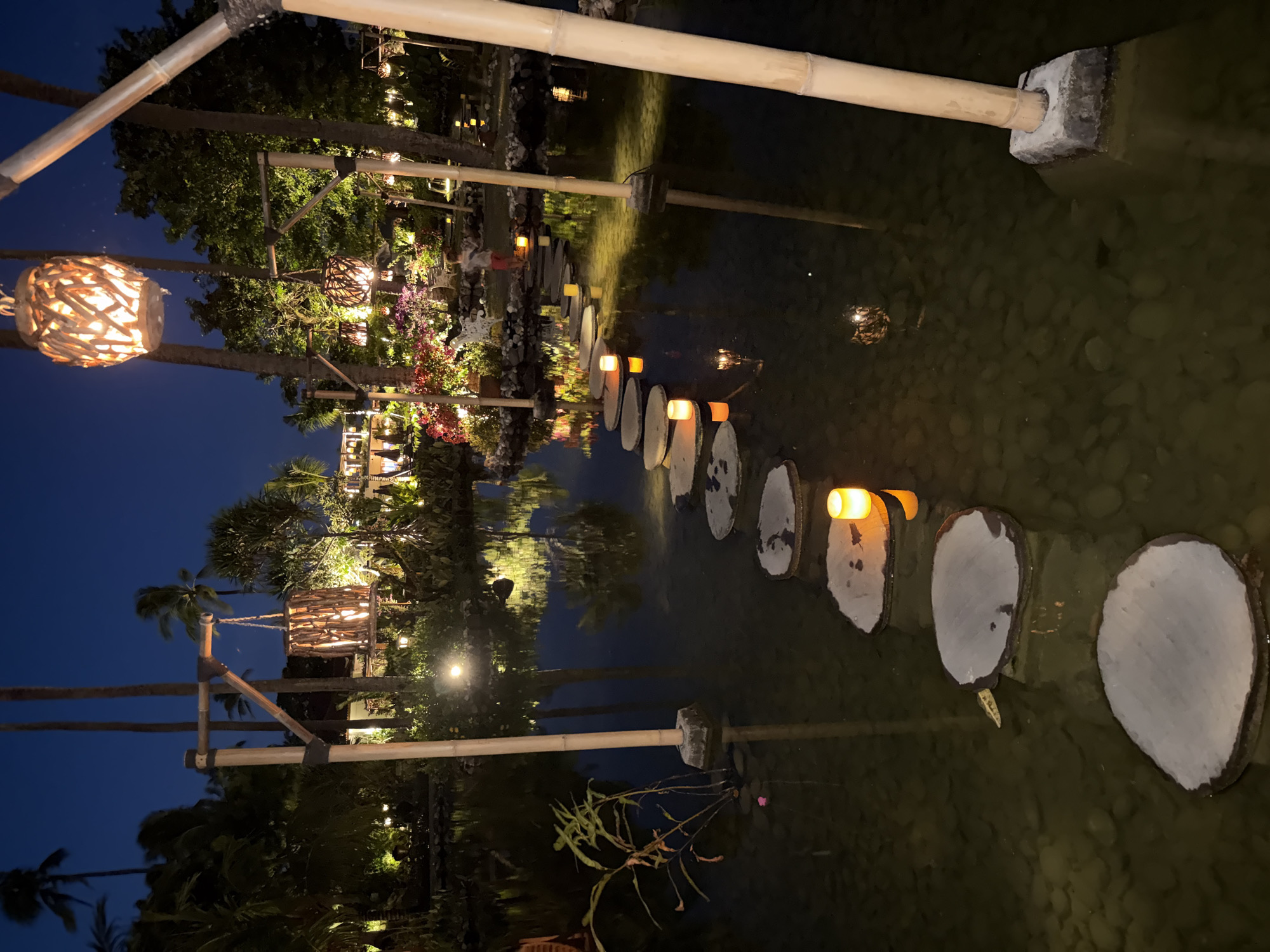 The image displays a serene, illuminated outdoor landscape at night, characteristic of a tropical resort or garden in Kuta Selatan, Indonesia. A shallow body of water, possibly a pond or decorative pool, dominates the scene, reflecting the surrounding lights and foliage.
A path of circular or oval stepping stones, appearing to be made of light-colored wood slices or concrete, crosses the water from the left to the right side of the frame. Each stepping stone is outlined in a darker material. Small, warm orange lanterns are strategically placed on or beside several of these stones, casting gentle glows onto the water and defining the pathway.
The water itself is dark and clear enough to reveal pebbles and stones on its bed, and its surface acts as a mirror, reflecting the numerous artificial lights and the lush vegetation. Surrounding the water are abundant tropical plants, including tall palm trees and dense green foliage, some adorned with visible red or pink blossoms in the background.
Decorative light fixtures, crafted from woven natural materials like wood or bamboo, hang from trees and overhead structures, providing additional warm illumination. A sturdy bamboo railing, supported by concrete anchors, extends across a section of the water. The sky above is a deep twilight blue, indicating a clear night. No people are visible, contributing to the tranquil and peaceful atmosphere. The scene evokes a sense of calm and inviting beauty, ideal for an evening stroll in a tropical setting. No visible text is present.