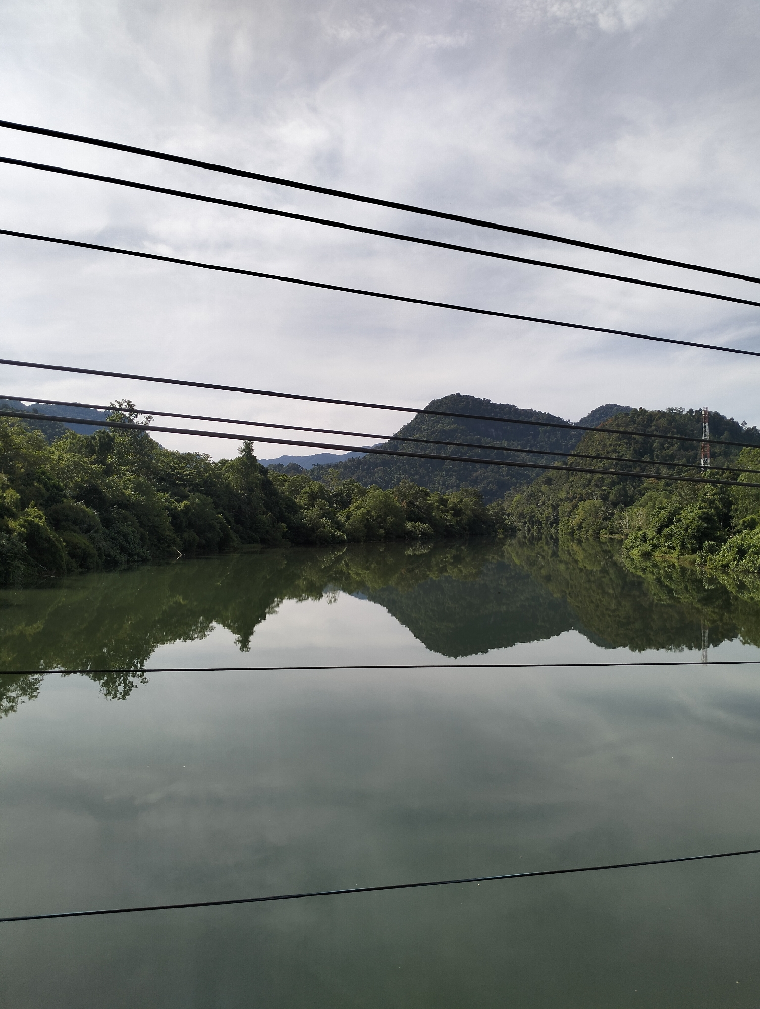 The image shows a landscape of a body of water surrounded by lush green hills and vegetation, likely a river or lake, in Lamseunia, Indonesia. The water's surface reflects the sky and the surrounding hills, creating a mirrored effect. Several black wires, possibly electrical or telephone, are strung across the top portion of the image. A tall, thin tower with red and white markings is visible on one of the hills in the background. The sky is overcast with a mottled pattern of clouds. No people are visible in the frame.
