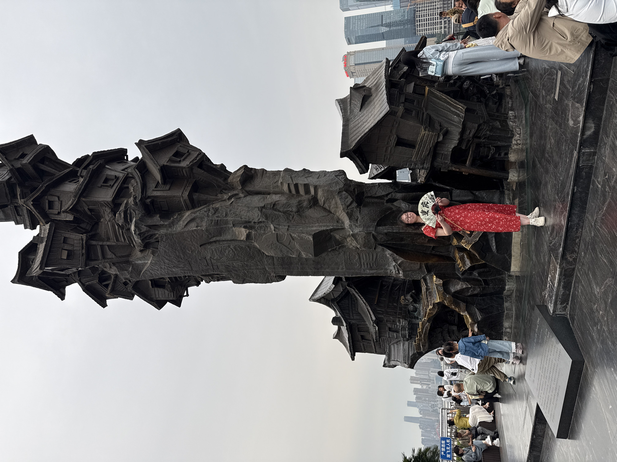 The image shows a tall, vertical sculpture resembling stacked, stylized houses, with a rocky, textured column beneath. The sculpture is made of a dark material, possibly bronze or stone. A woman in a red floral dress and white sneakers stands in front of the sculpture, holding a white fan with writing on it. Other people are present around the sculpture, some standing and some sitting. In the background, there's a cityscape with tall buildings visible through a slightly overcast sky. The location is the city of Yuzhong District, China.