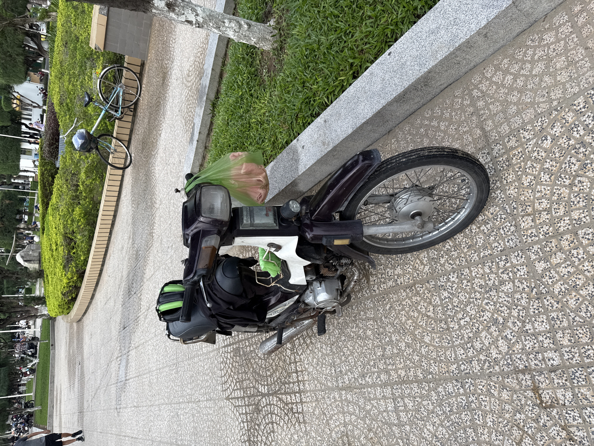 The image shows a parked motorcycle in a park in Nha Trang, Vietnam. The motorcycle is dark purple with a black seat and saddlebags. It has a green helmet on the seat and a green plastic bag hanging from the handlebars. A light blue bicycle is parked behind the motorcycle. The park has a paved walkway, a grassy area, and trees. In the background, there are buildings and other people walking. The overall impression is one of a typical urban park scene.