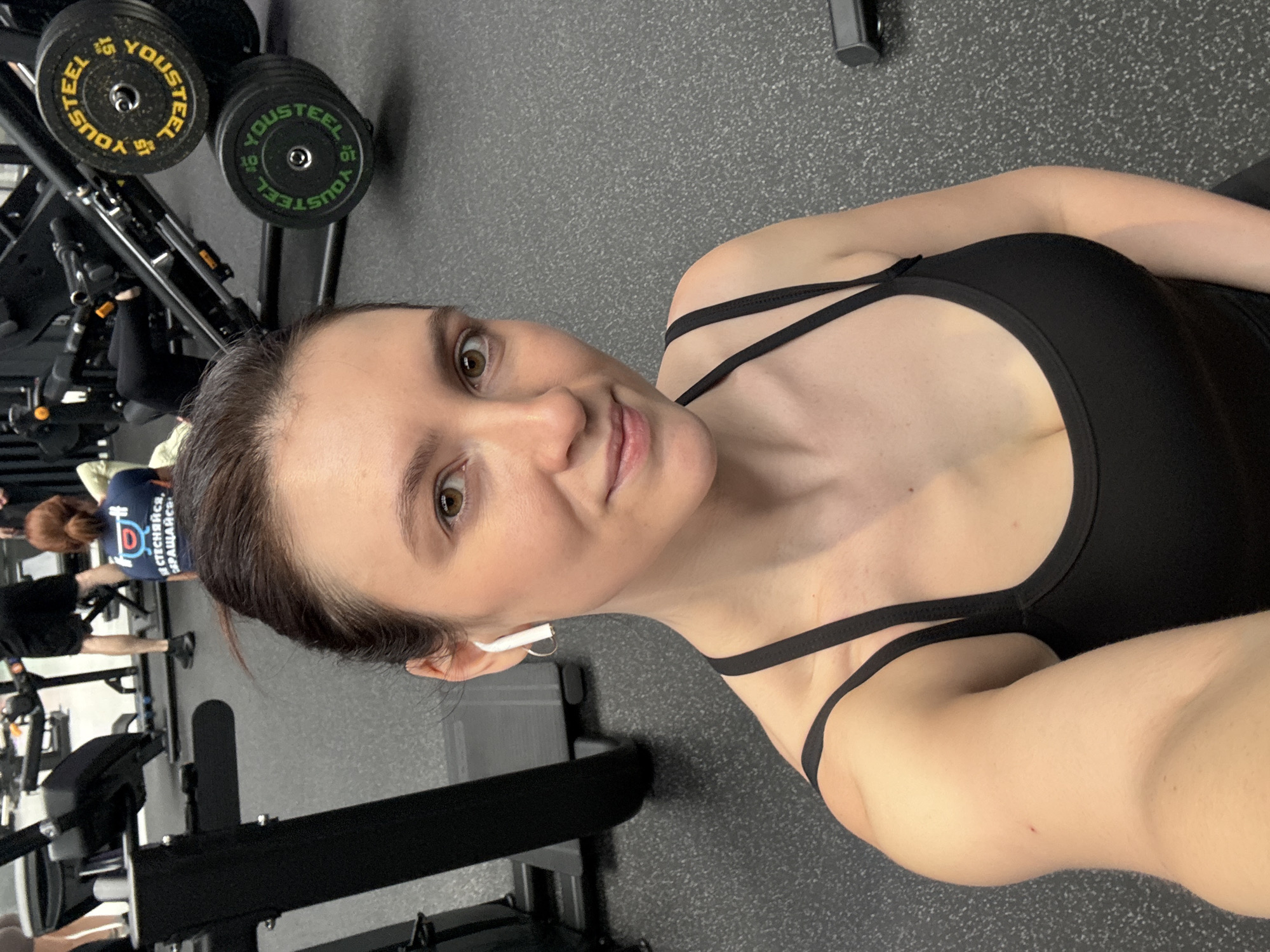 The image shows a woman in a black sports bra at a gym. She is taking a selfie, with gym equipment visible in the background. There are weight plates labeled "Yousteel." Other people are present in the background, engaged in workouts. The location is consistent with a gym setting. There is no evidence to confirm the city of Kazan, Russia.