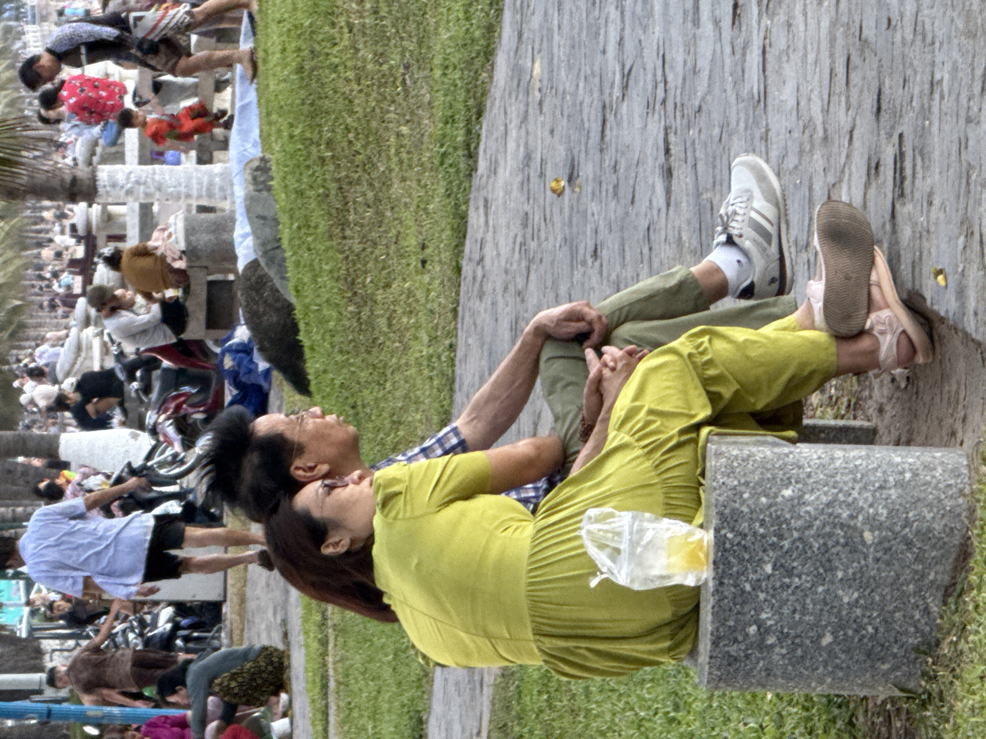 In Nha Trang Ward, Vietnam, two people are resting on a stone bench by a paved walkway, with a grassy area beside them. The man in the foreground is wearing a blue plaid shirt and khaki pants, and is leaning back, resting his head on his hand. Beside him, a woman in a bright yellow dress is also leaning back, with her head tilted up. Her right arm is resting on the man's leg. A clear plastic bag with a yellow object inside is tucked beside her. The image has a shallow depth of field, with the background out of focus. Further in the background, other people are visible walking and sitting, suggesting a public park or promenade. The scene appears to be set during the daytime, possibly late morning or early afternoon given the lighting. The overall mood seems relaxed and casual, with the two individuals enjoying a moment of repose.