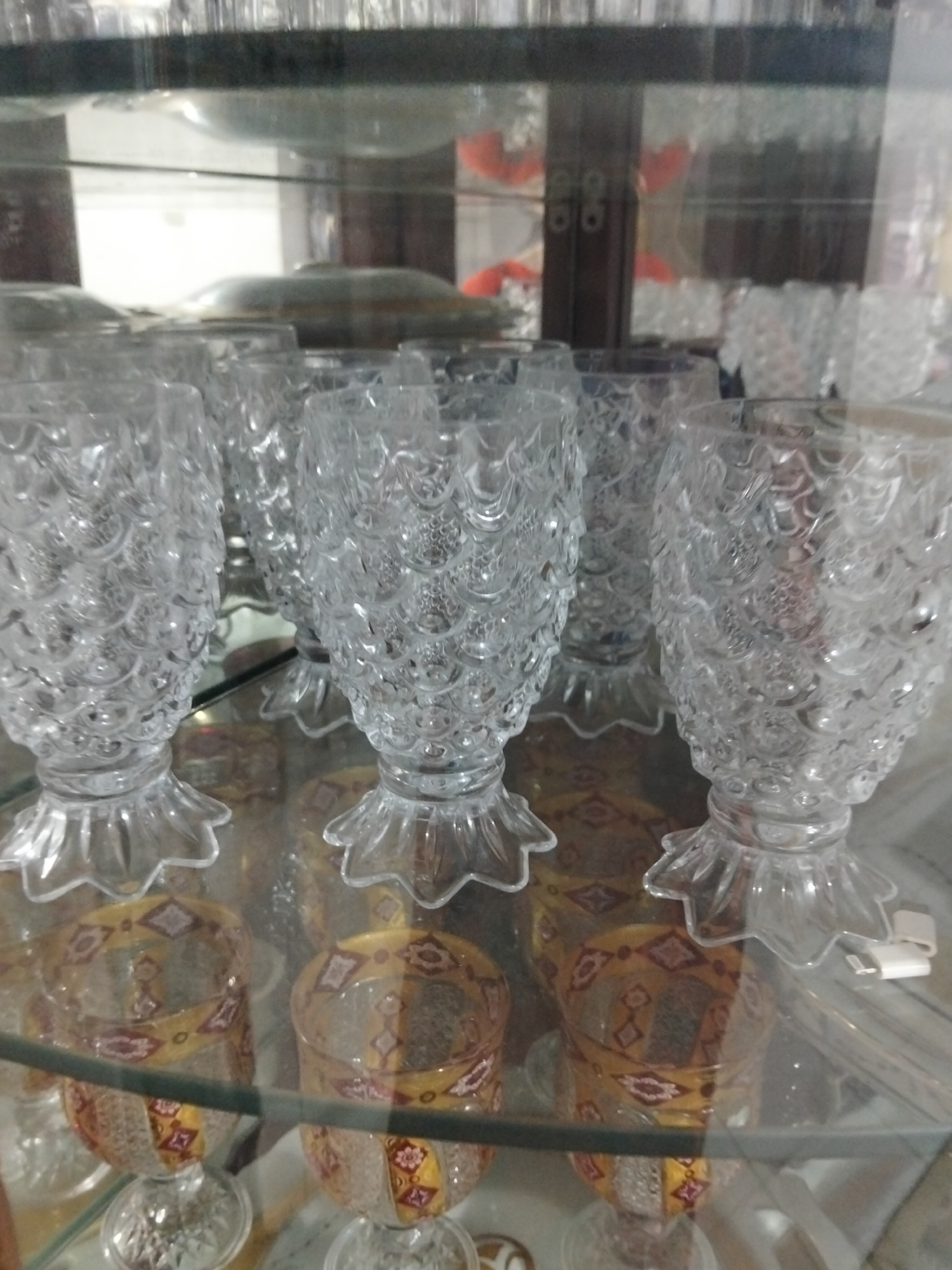 In Payagaboh, Indonesia, a glass cabinet displays a collection of glassware. The cabinet has multiple shelves, with the front-most shelf showing several clear glass cups with intricate, scale-like textures and fluted bases. Some of these cups are positioned above smaller, decorative glasses that feature a yellow and red diamond pattern. Reflections are visible on the glass surfaces, suggesting an indoor setting. The lighting appears to be ambient, casting soft shadows.