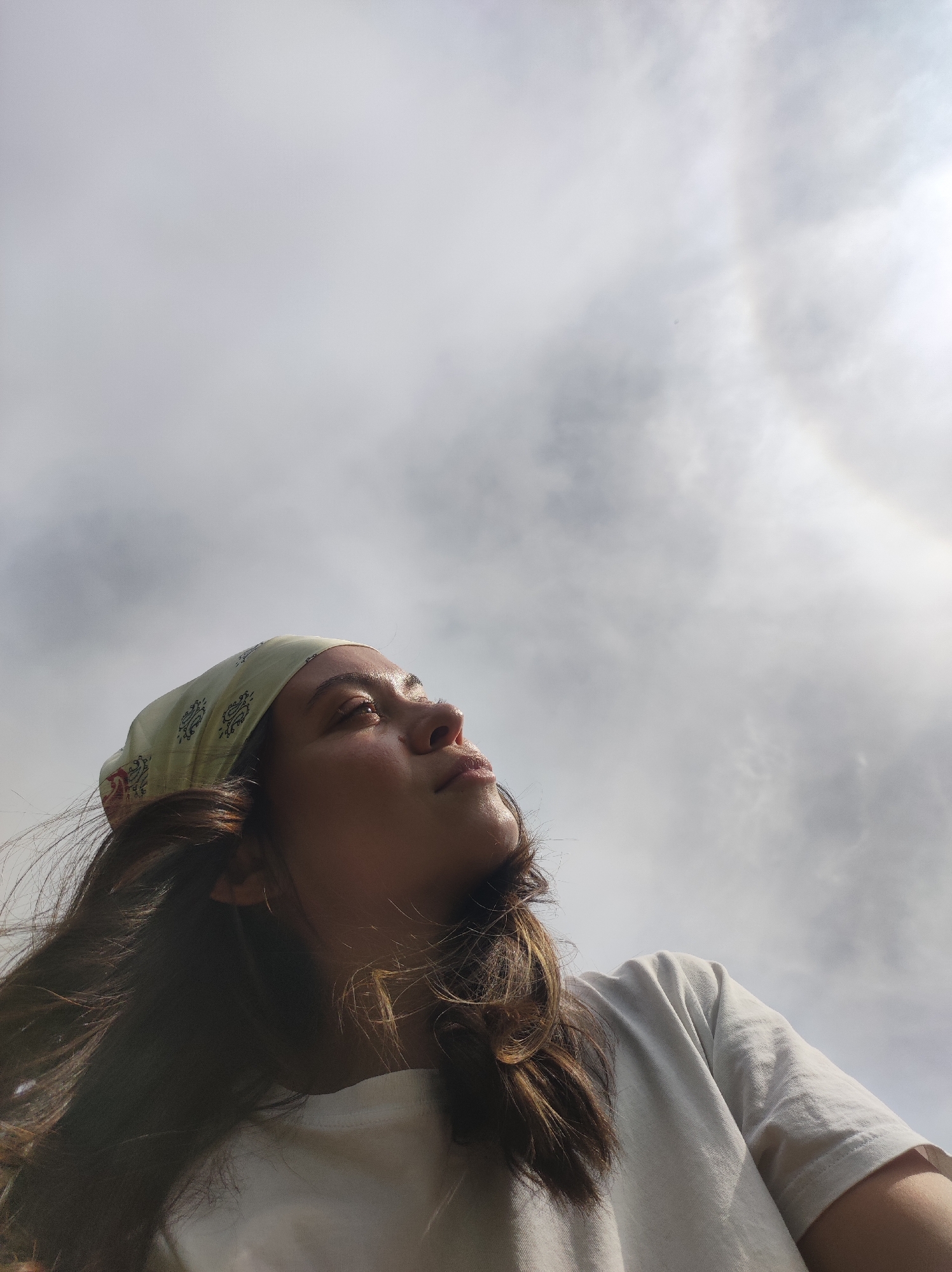 A woman with long brown hair and a light-colored bandana is looking upward. She is wearing a white t-shirt. The background is a cloudy sky with a halo around the sun. The location cannot be determined.