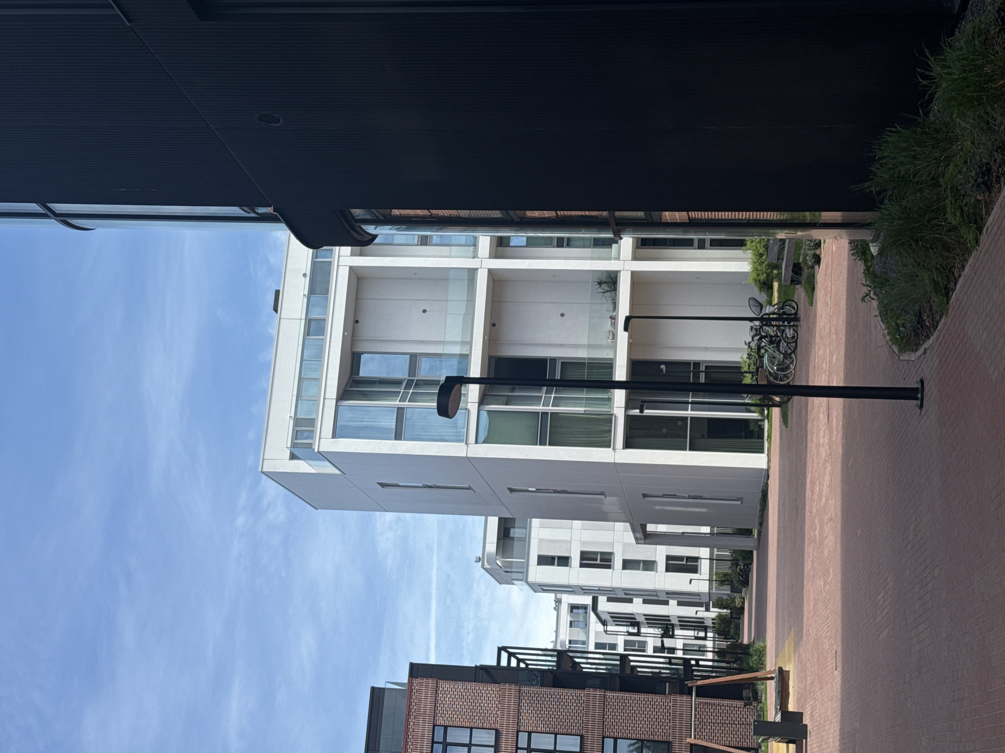 This is a vertical, eye-level shot of a modern courtyard in Tallinn, Estonia, on a sunny day with a few wispy clouds in the bright blue sky. The foreground is dominated by a red brick paved walkway that curves to the right. To the left, a section of a red brick building with balconies and dark-framed windows is visible. In the middle ground, a tall, white, modern apartment building with large glass windows and balconies rises, featuring a geometric design. Adjacent to it, other similar white buildings recede into the distance. A black lamppost with a modern design stands in the middle of the walkway. In the mid-ground, to the left of the white building, a set of bicycles are parked near a wooden bench. To the right, a dark, corrugated building with a curved glass section partially obscures the view of the white buildings. Green bushes and grass are visible along the edges of the walkway and around the base of the buildings.
