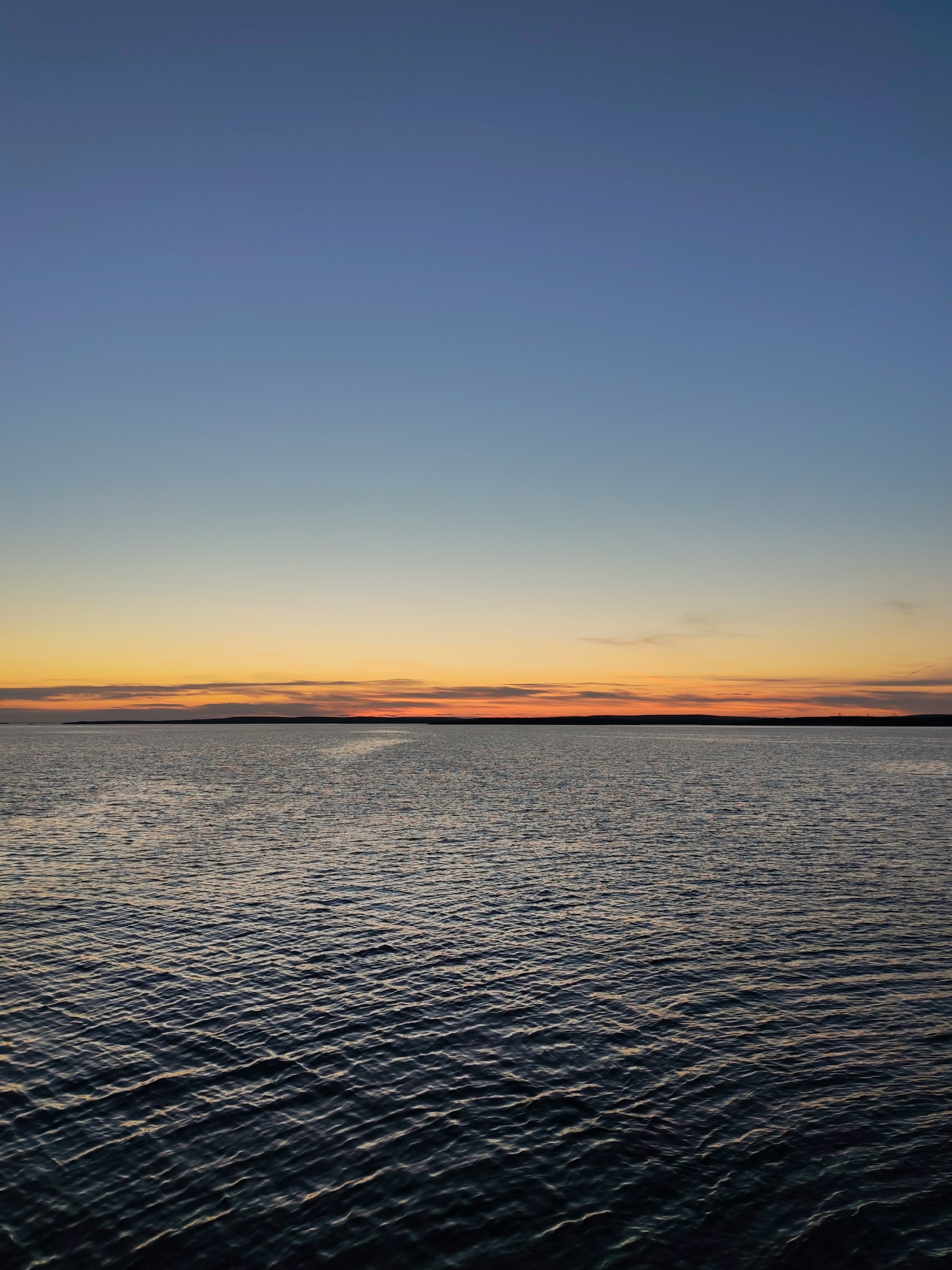 The image depicts a serene body of water under a gradient sky, transitioning from a deep blue at the top to warm oranges and yellows near the horizon. The water's surface is rippled, suggesting a gentle breeze. A dark, silhouetted landmass is visible in the distance, marking the horizon line. The sky is clear with subtle cloud formations that catch the fading sunlight. The scene appears to be a natural landscape, likely a coastal area or a large lake. There are no people, animals, buildings, or man-made objects clearly visible that would indicate a specific urban location like Shal'skii, Russia. The overall impression is one of natural beauty and tranquility at dusk or dawn.