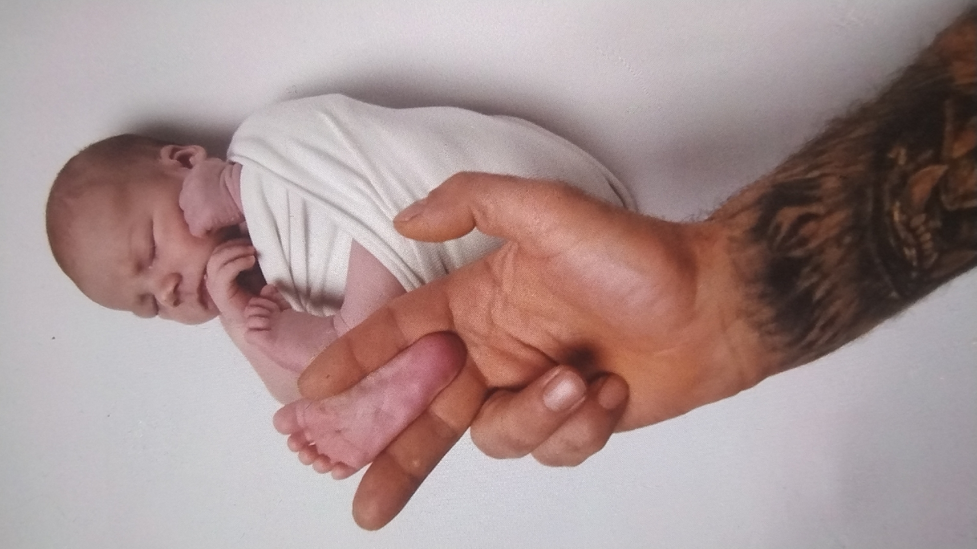 A newborn baby, swaddled in white fabric, is held by a large adult hand. The baby is asleep with its eyes closed and its face resting on its hands. The baby's feet are visible and appear to be pink. The adult hand is tanned and has tattoos on the forearm, which appear to be animalistic in nature. The background is a plain white wall. There is no information about the location in the city of Mykolaiv, Ukraine.