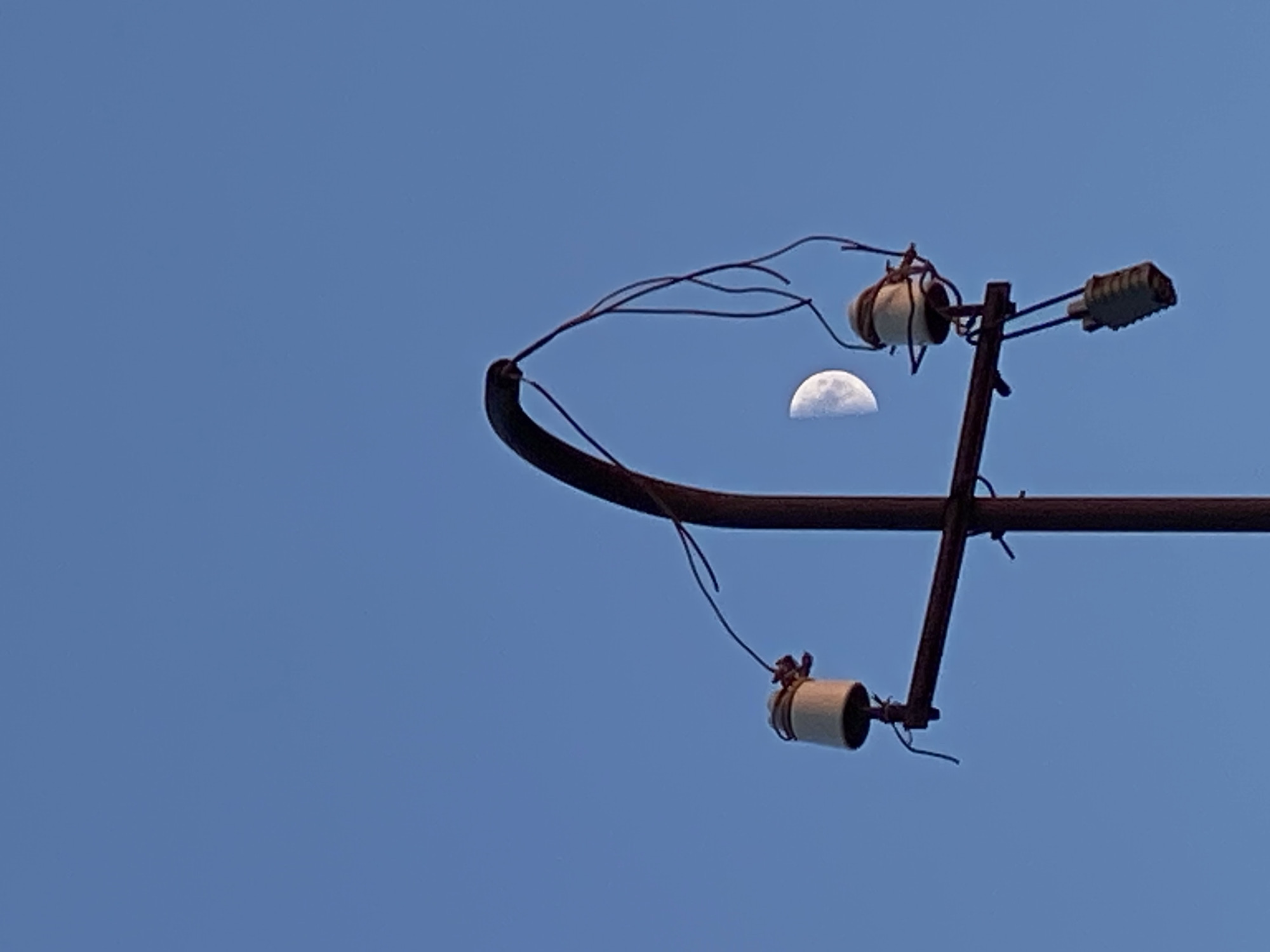 A close-up, low-angle, full shot shows a utility pole against a clear, bright blue sky. The pole is made of dark brown metal, with a curved upper section. Attached to the pole is a horizontal crossarm, also dark brown, from which hang two ceramic insulators. The insulators are white with brown caps, and wires are attached to them, some of which are loose and dangling. One insulator has a grey, ribbed object hanging from it. To the right of the pole, in the middle ground of the image, is a crescent moon, partially obscured by the wires. The scene is set in Cairaclia, Moldova.