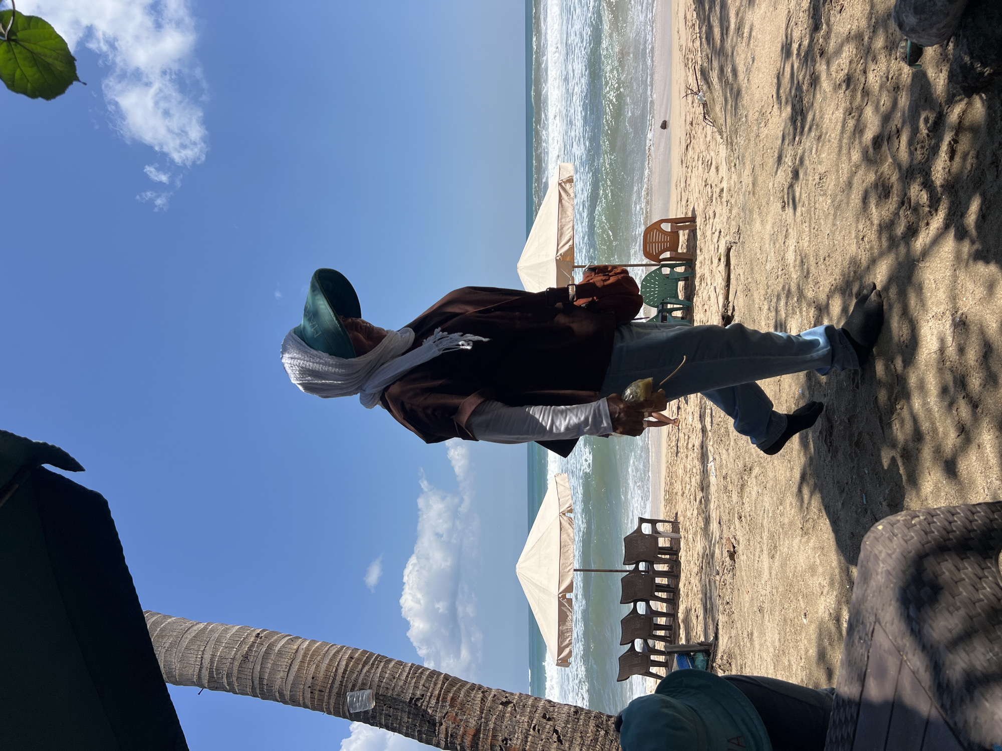 A woman walks on a beach in Kuta, Indonesia, carrying a small object in her hand. She is wearing a light-colored headscarf, a dark brown long-sleeved shirt, and light blue pants. She also has a green sun hat and a small brown bag slung over her shoulder. The beach is sandy, with several plastic chairs and umbrellas set up in the background. The ocean is visible, with waves rolling in. The sky is blue with some white clouds. The scene is lit by the sun, creating shadows on the sand.