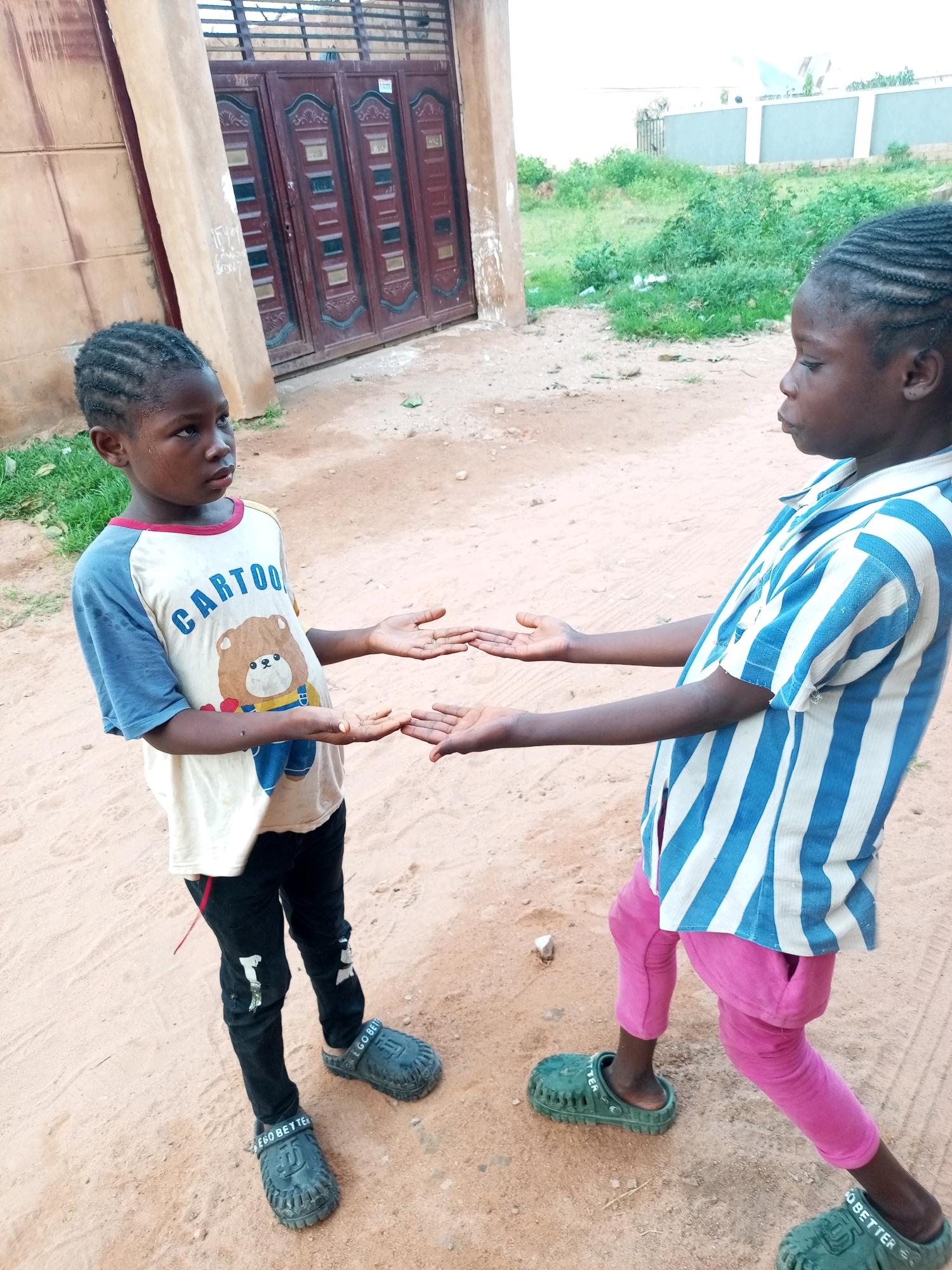 Two young children, one appearing to be a girl and the other a boy, stand facing each other outdoors in what appears to be a residential area in Tempure, Nigeria. The child on the left, wearing a t-shirt with a cartoon bear and the word "CARTOON" printed on it, black pants, and dark clogs, holds their hands open, palms facing upwards. The child on the right, wearing a blue and white striped shirt and pink pants, also holds their hands open, palms facing upwards, in a gesture that suggests they are interacting. The ground is sandy and there is some sparse vegetation in the background. A large, decorative gate is visible behind the child on the left, and a light-colored wall and more vegetation are behind the child on the right. The lighting suggests it is daytime.