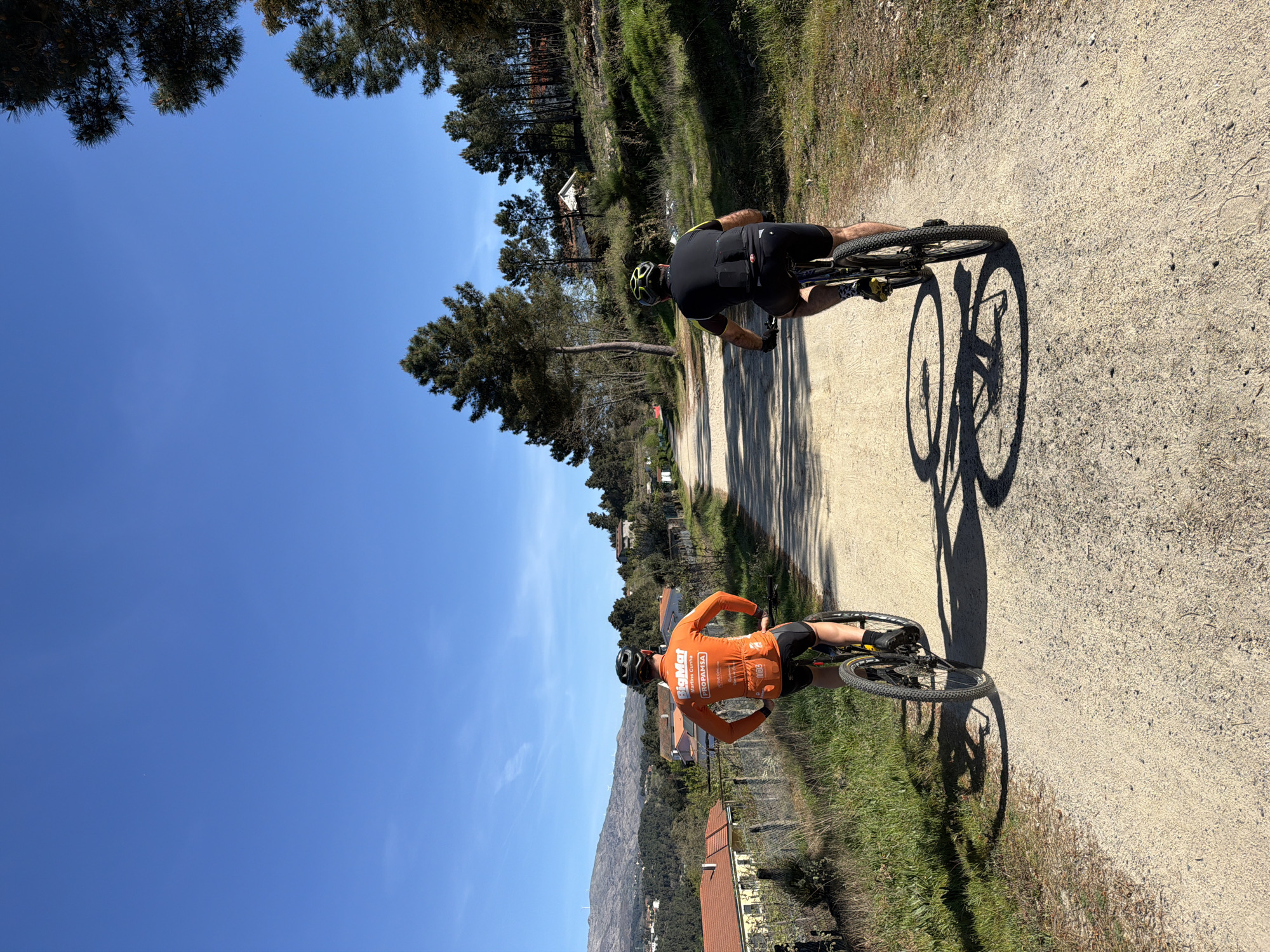 Two cyclists ride uphill on a dirt path. The cyclist in the foreground wears an orange jersey with the word "BigMat" and "Martin Cunha" printed on it, while the cyclist behind him wears a black jersey. The scene takes place in Vila Real, Portugal, as suggested by the architecture and the general landscape. The environment features trees lining the path, and a blue sky with sparse clouds. In the distance, a mountain range is visible. The cyclists' shadows are cast on the path.