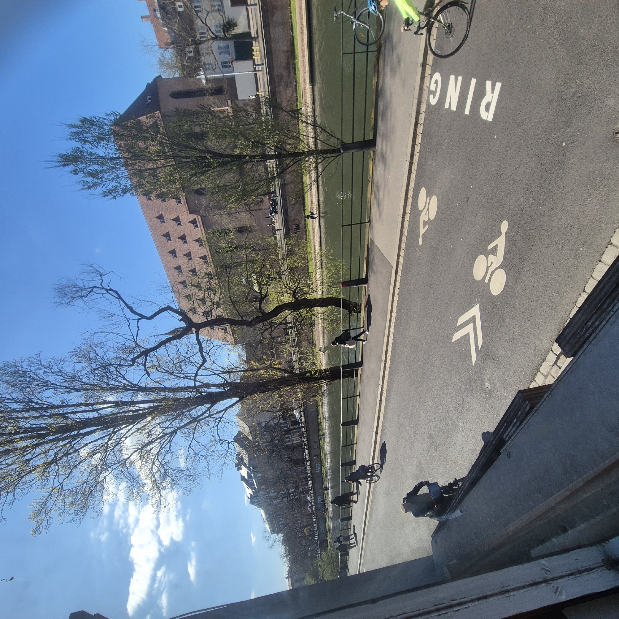 The image depicts a riverside pathway in Strasbourg, France, marked for bicycle use with the word "RING" and a bicycle symbol painted on the asphalt. A canal runs parallel to the path, separated by a low railing. Several people are visible on the pathway, some walking and at least one riding a bicycle. The architecture suggests a historical European city with brick and stone buildings lining the opposite side of the canal. Bare trees are scattered along the waterway, reaching towards a blue sky with scattered clouds. The perspective appears to be from an elevated position, perhaps a window or bridge.