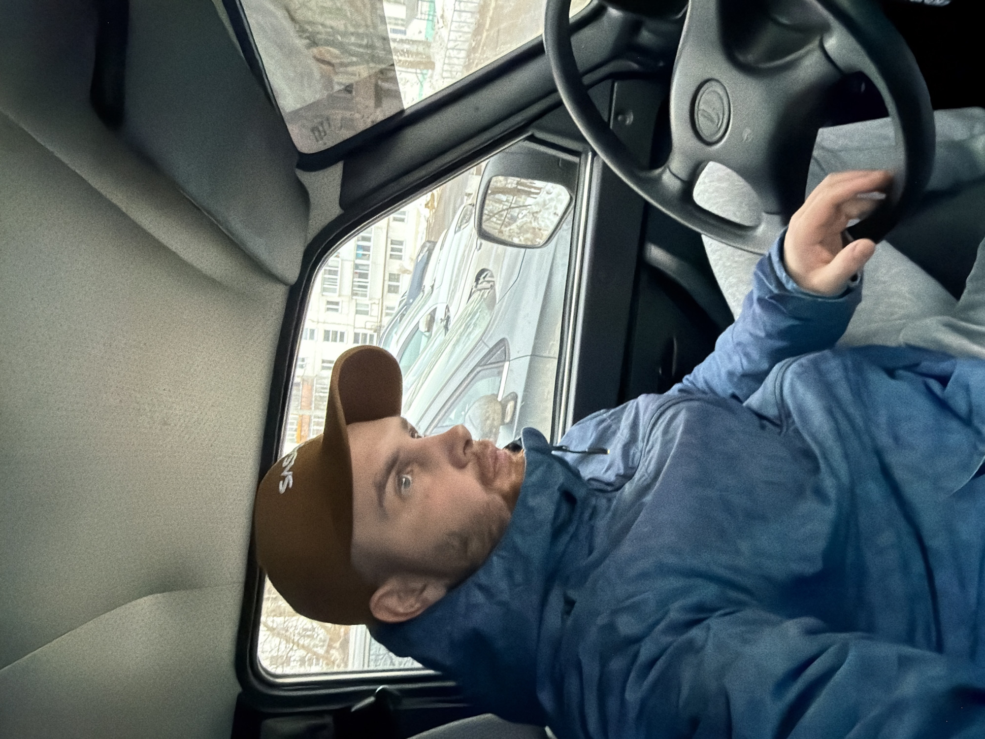 A man is seen inside a car, wearing a brown cap and a blue jacket. He appears to be in the driver's seat, with a steering wheel visible in front of him. Through the car window, a cityscape is visible, identified as Chaikovsky, Russia, based on the building architecture and urban landscape. The man is looking out of the window. The interior of the car is visible.