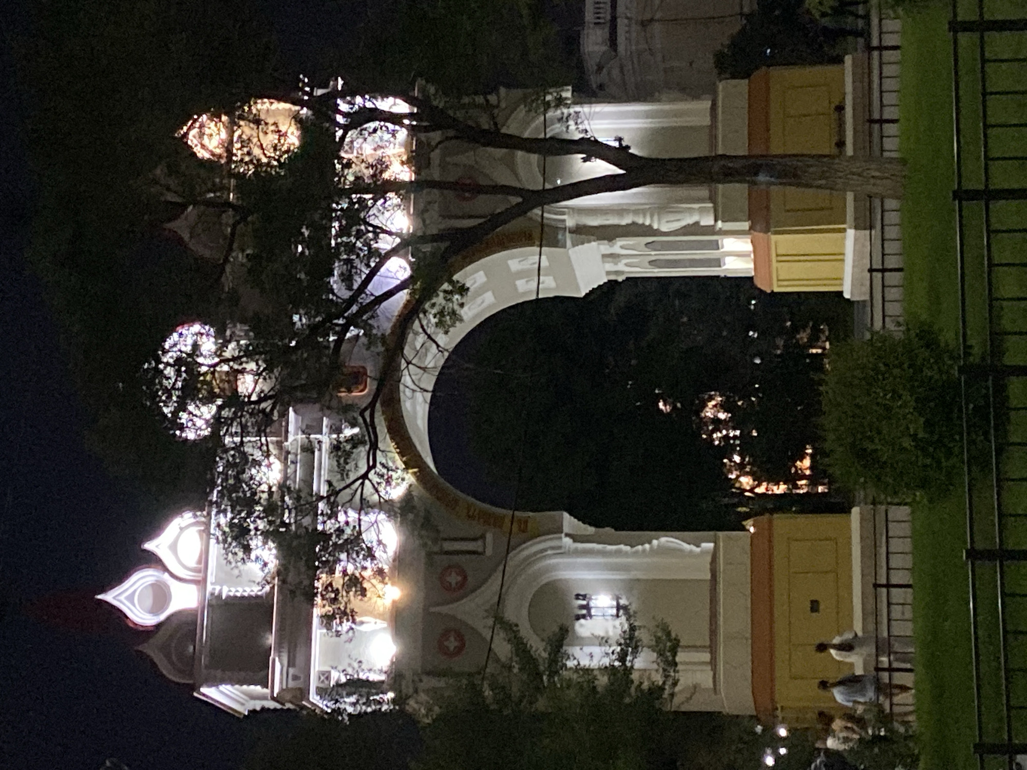 The image captures the Amur Oblast Government Building, also known as the "Arch of the Amur," in Blagoveshchensk, Russia, at night. The building features a prominent archway and is illuminated with bright lights, highlighting its architectural details. Several domed structures are visible on top. A tree partially obscures the right side of the arch. In the foreground, a black metal fence encloses a grassy area, and several people are seen standing and walking near the base of the building, in front of a yellow facade. The overall scene depicts an illuminated monument in an urban setting at night.